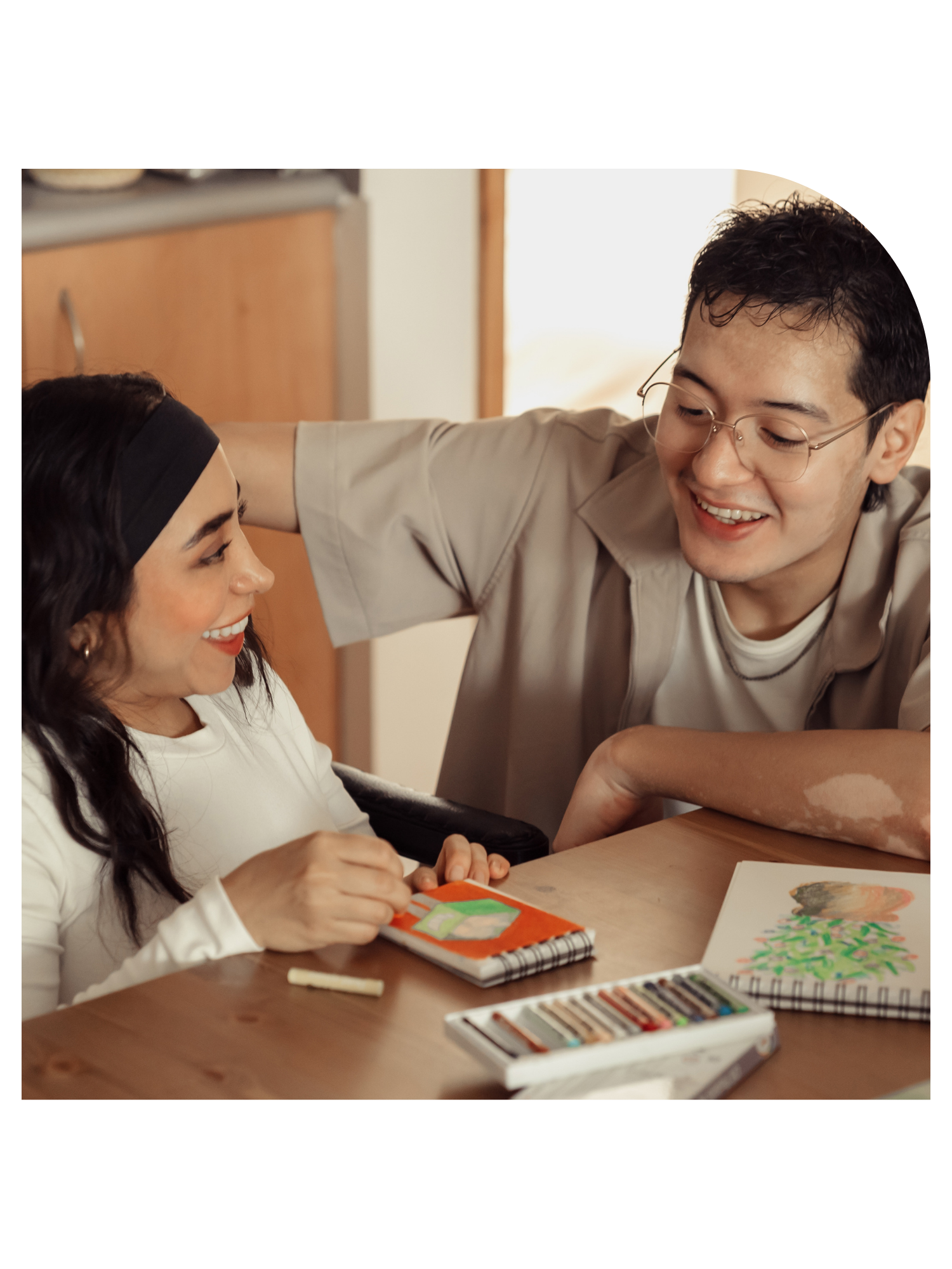 A man with glasses smiling and looking at a woman wearing a black headband, who is engaged in a creative activity at a wooden table, surrounded by art supplies and drawings.