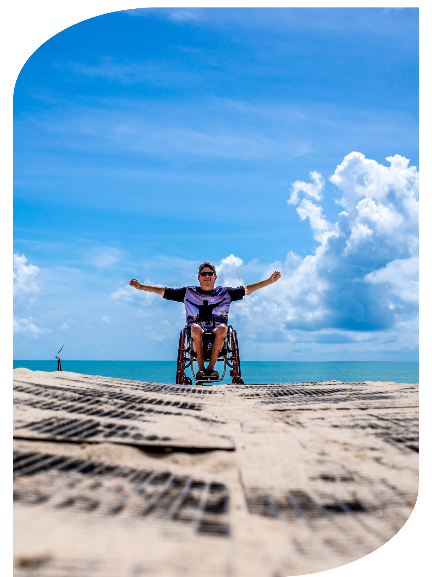 A man in a wheelchair at the beach, smiling with arms outstretched under a blue sky with scattered clouds.