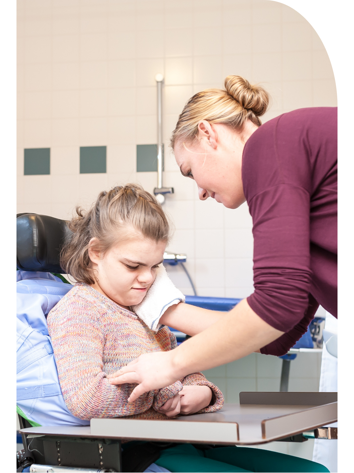 A young girl in a wheelchair with A woman, likely a healthcare worker, giving attentive care to the girl.
