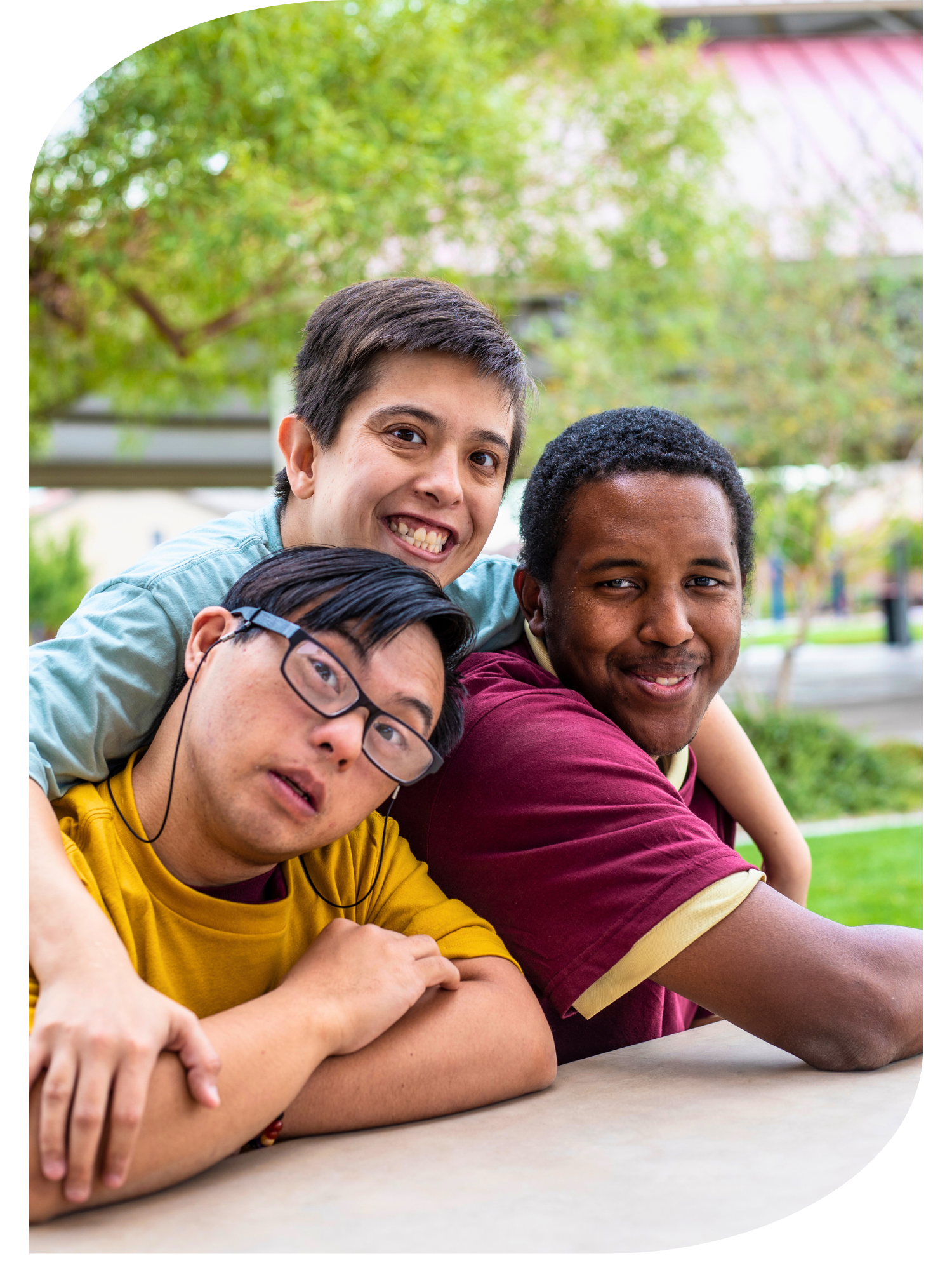 A group of three diverse young men sitting at a table outdoors in a park, smiling and leaning on each other.