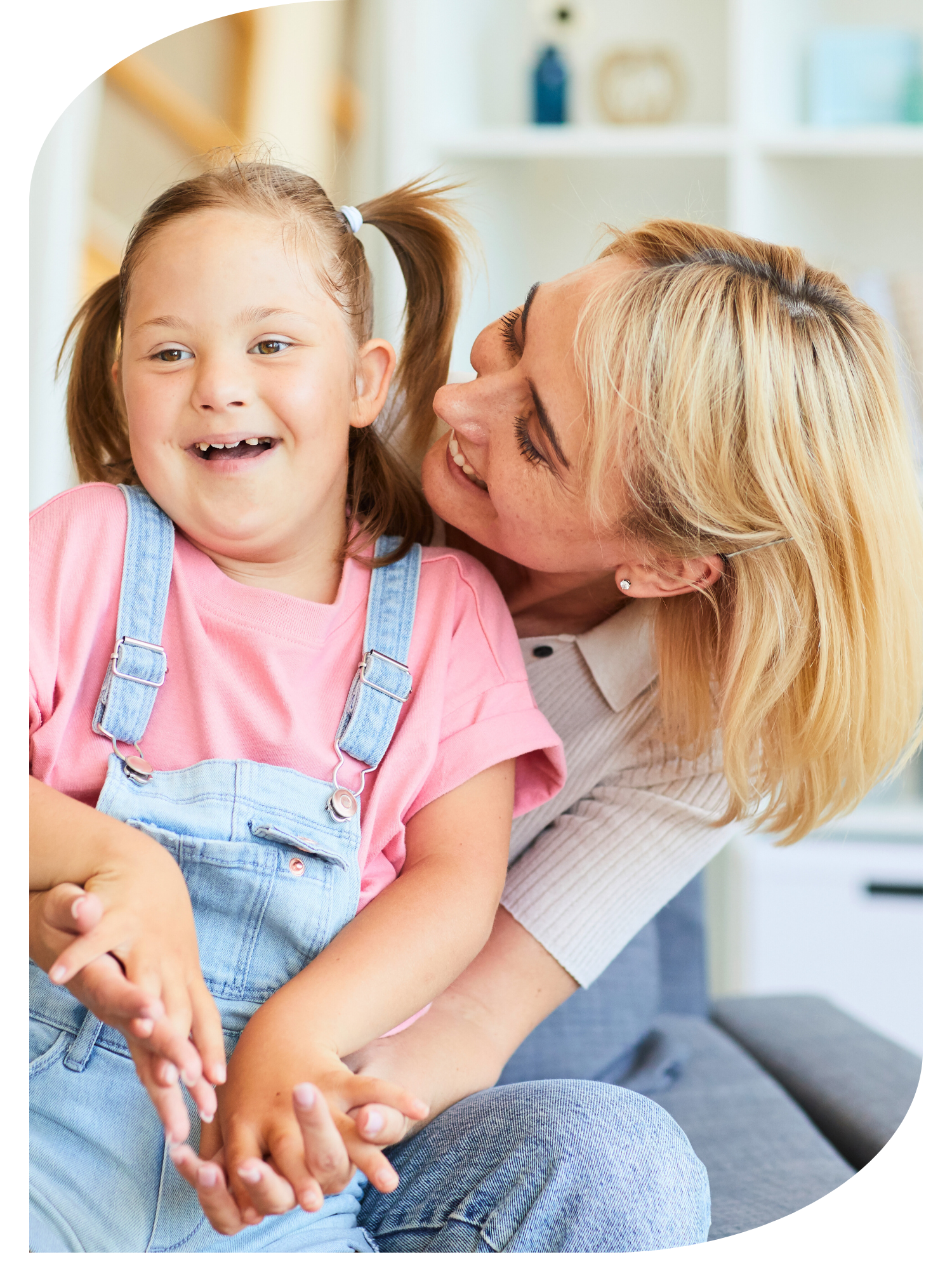 A young girl with brown hair in pigtails, wearing a pink shirt and denim overalls, smiling as she looks to the side while being embraced by an adult woman with blonde hair. They are in a bright room with shelves and decorative items in the background.