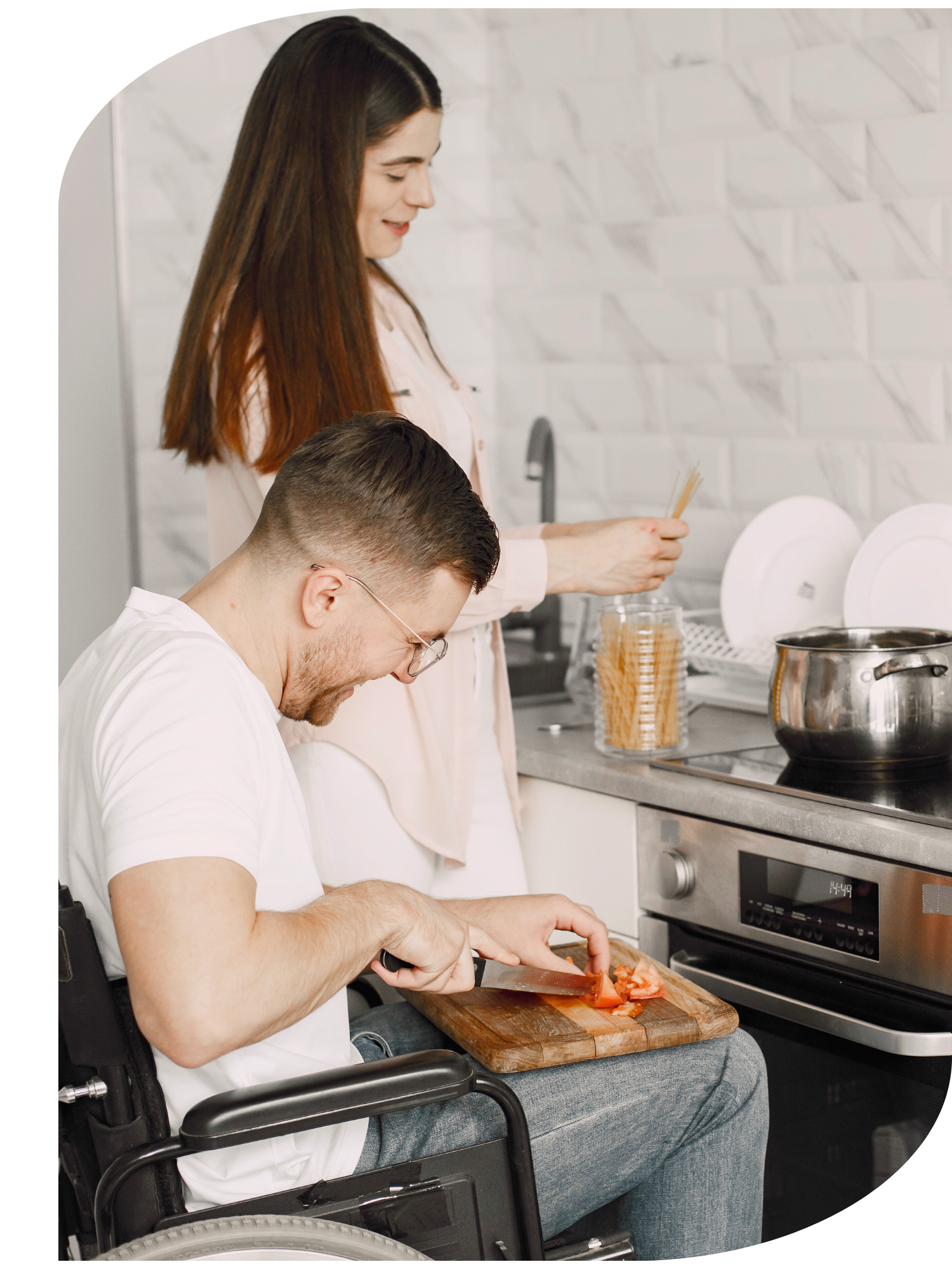 A young man in a wheelchair chopping tomatoes on a cutting board in a kitchen. A woman stands behind him,  preparing food near a stove and kitchen counter, with dishes and utensils visible.