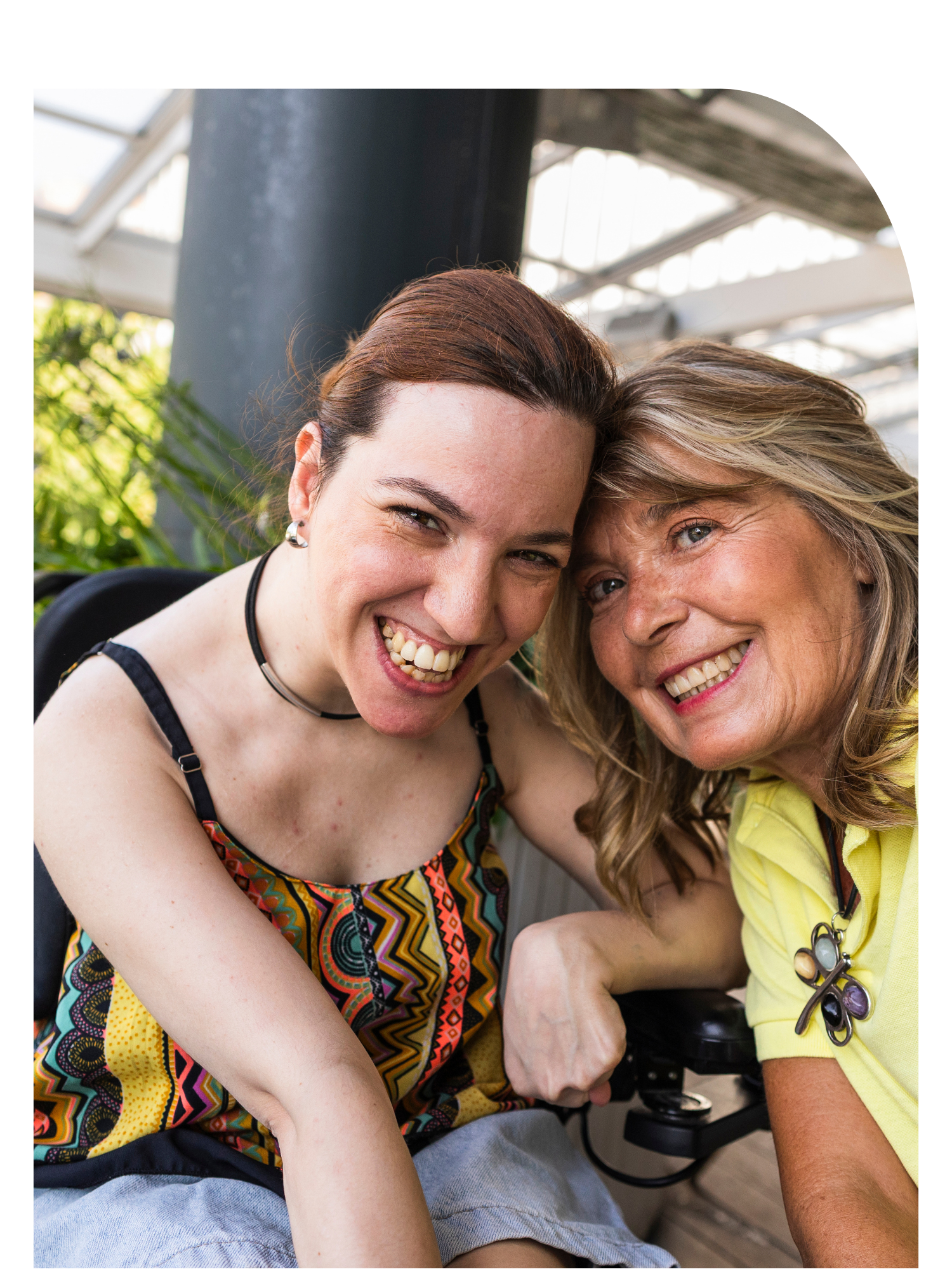 Two smiling women, one with red hair and the other with blonde hair, close together, showing their teeth, outdoors with a glass ceiling and green plants in the background.