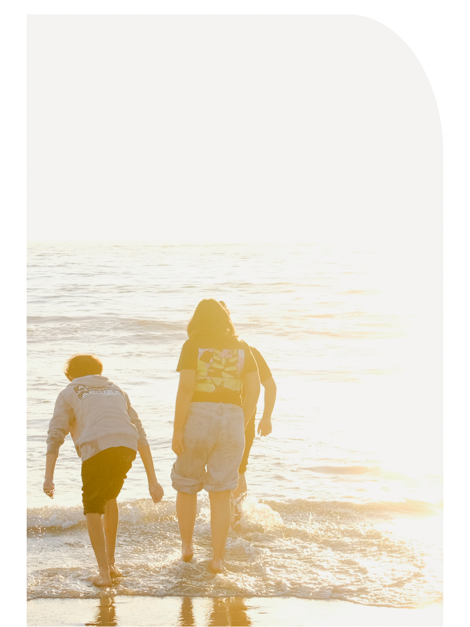 Three people at the beach during sunset, with two visible in the foreground and one further behind, all walking along the shoreline with the ocean and the setting sun in the background.