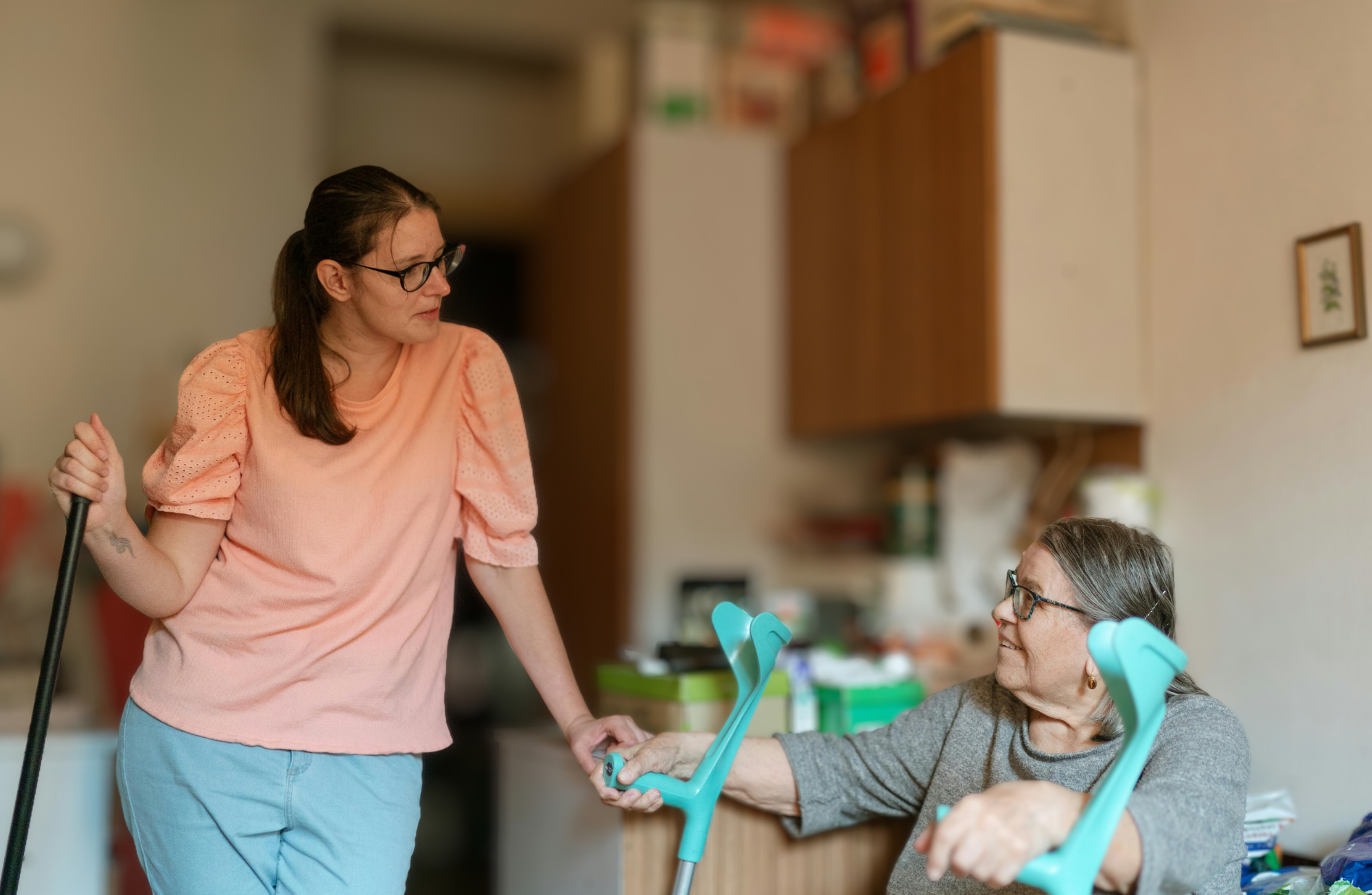 A woman wearing glasses and a peach-colored blouse stands next to an elderly woman with glasses, who is seated, both smiling as the seated woman holds hands with the standing woman in a room with kitchen cabinets and framed pictures on the wall.