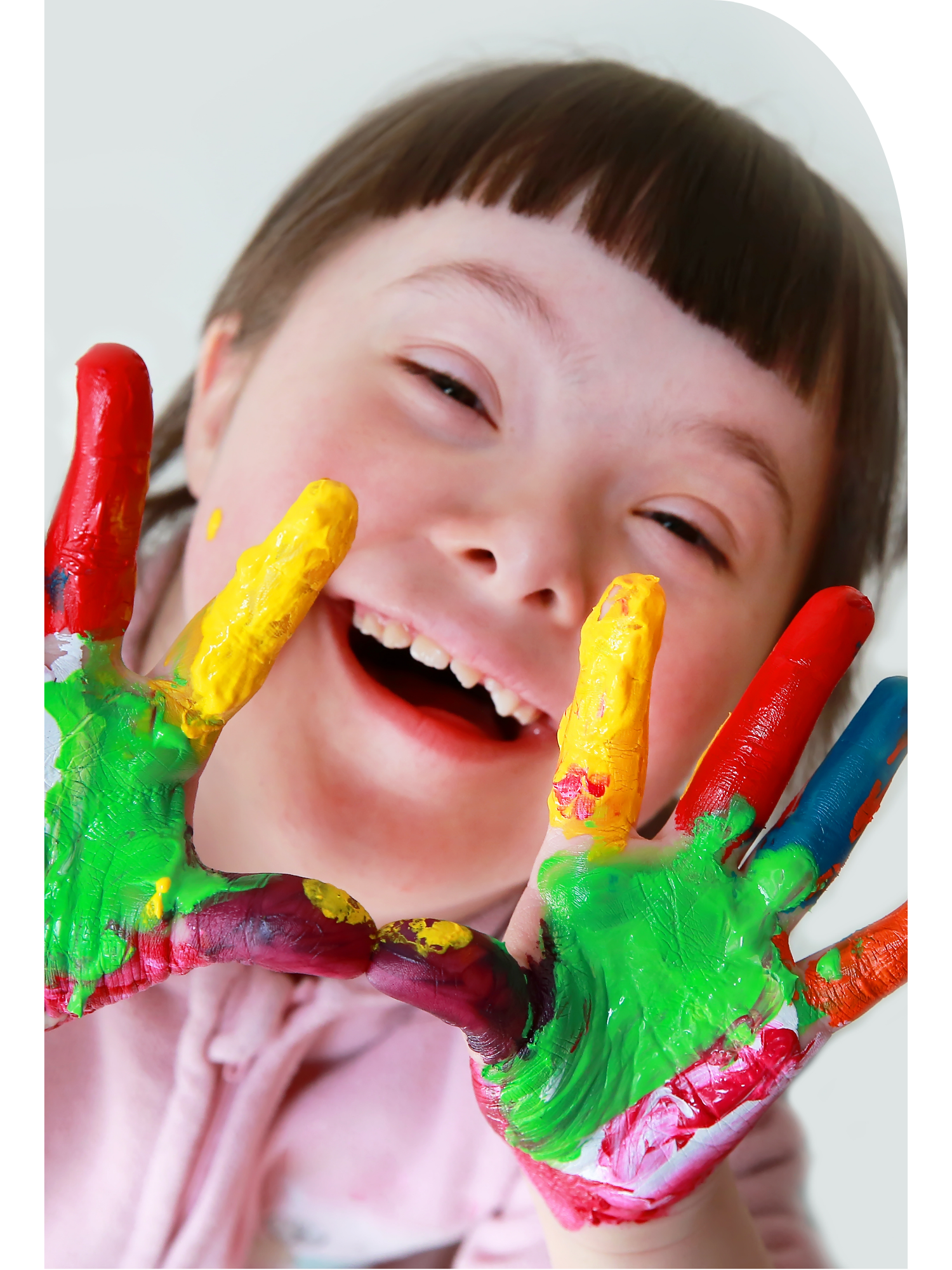Close-up of a young girl smiling with her eyes closed, wearing a light pink hoodie, holding her hand covered in colorful painted handprints up to her face.