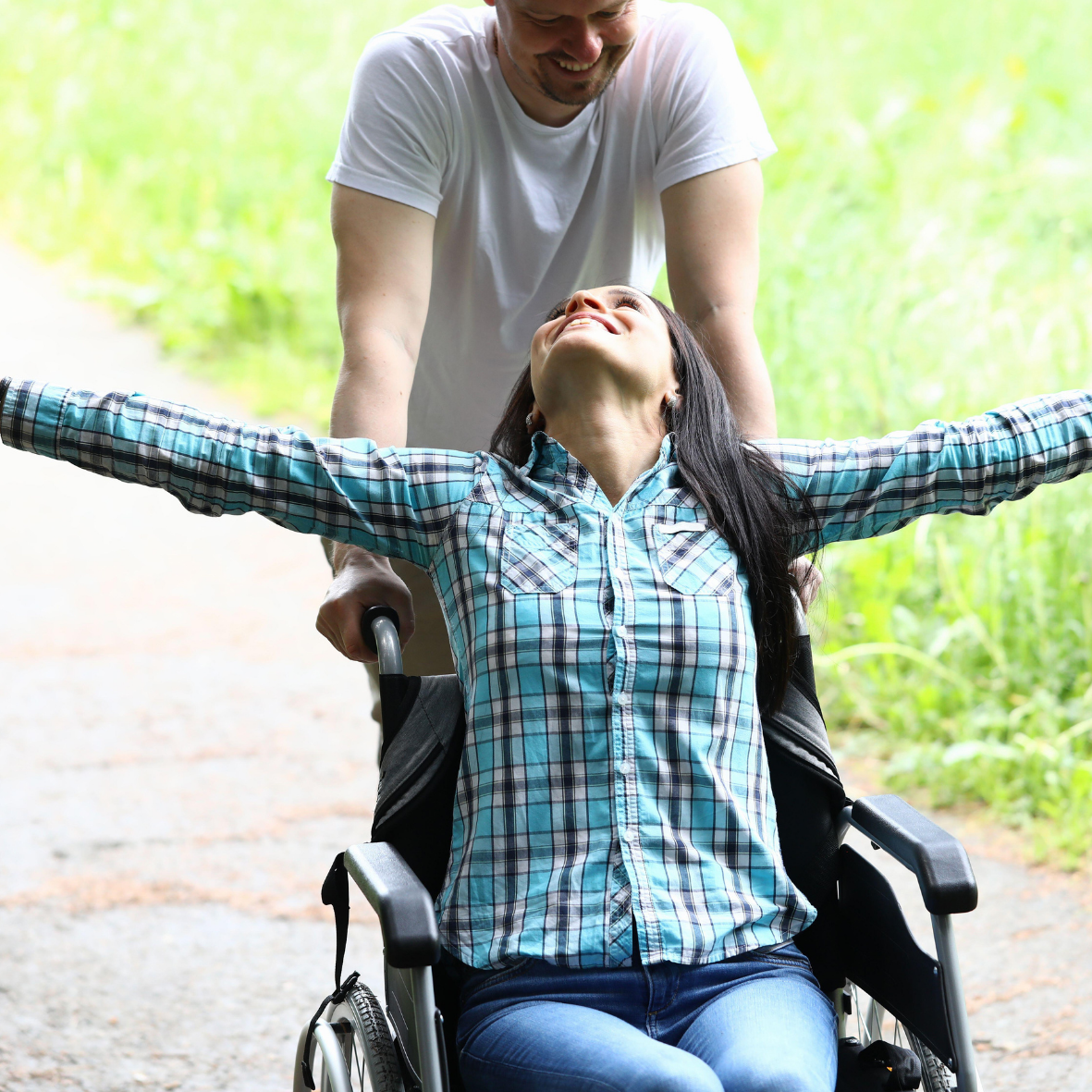 A woman in a wheelchair with arms outstretched, smiling, while a man pushes her on a path.