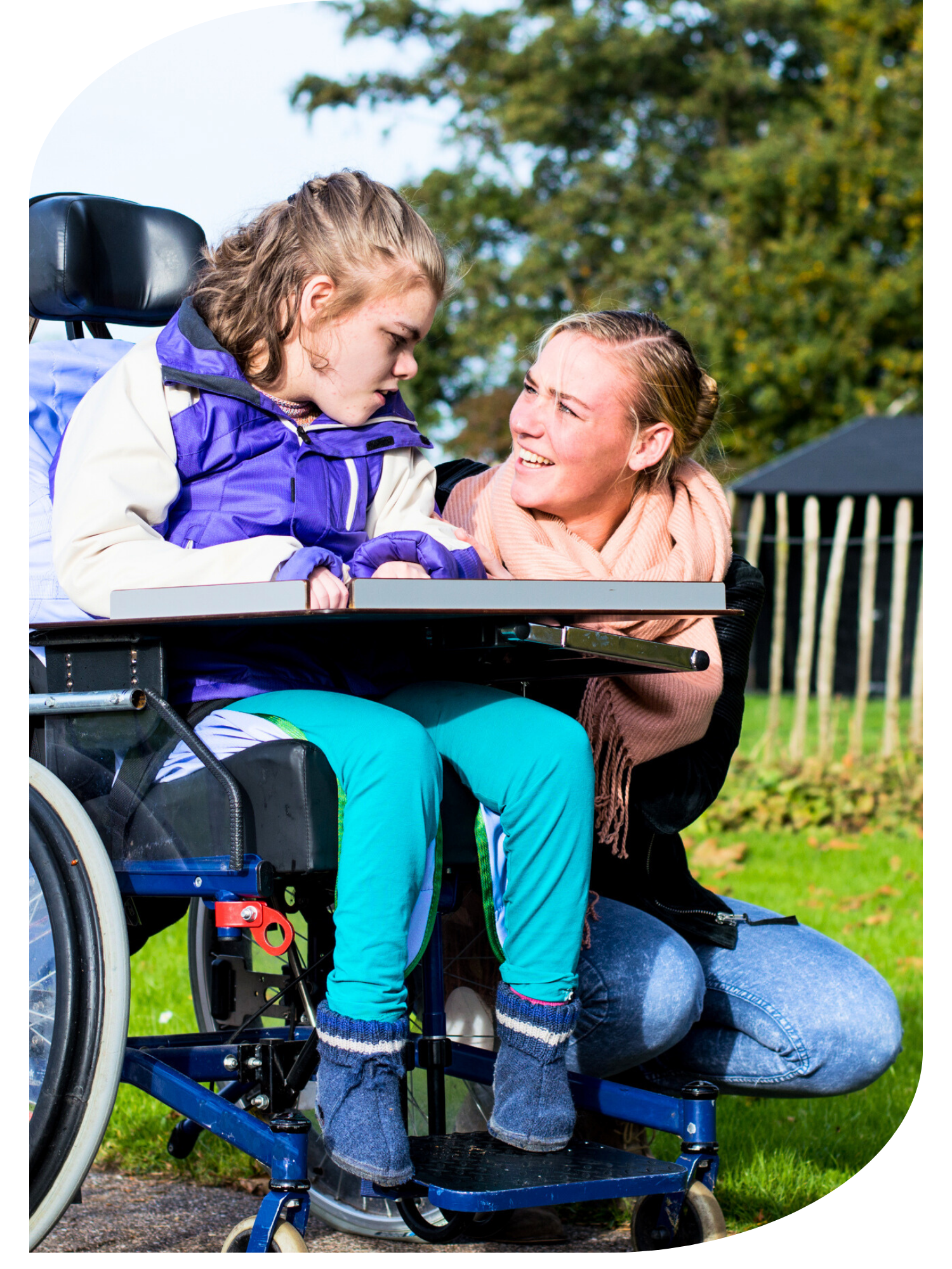 A young girl in a wheelchair and a woman outdoors in a park with green trees, smiling and looking at each other.