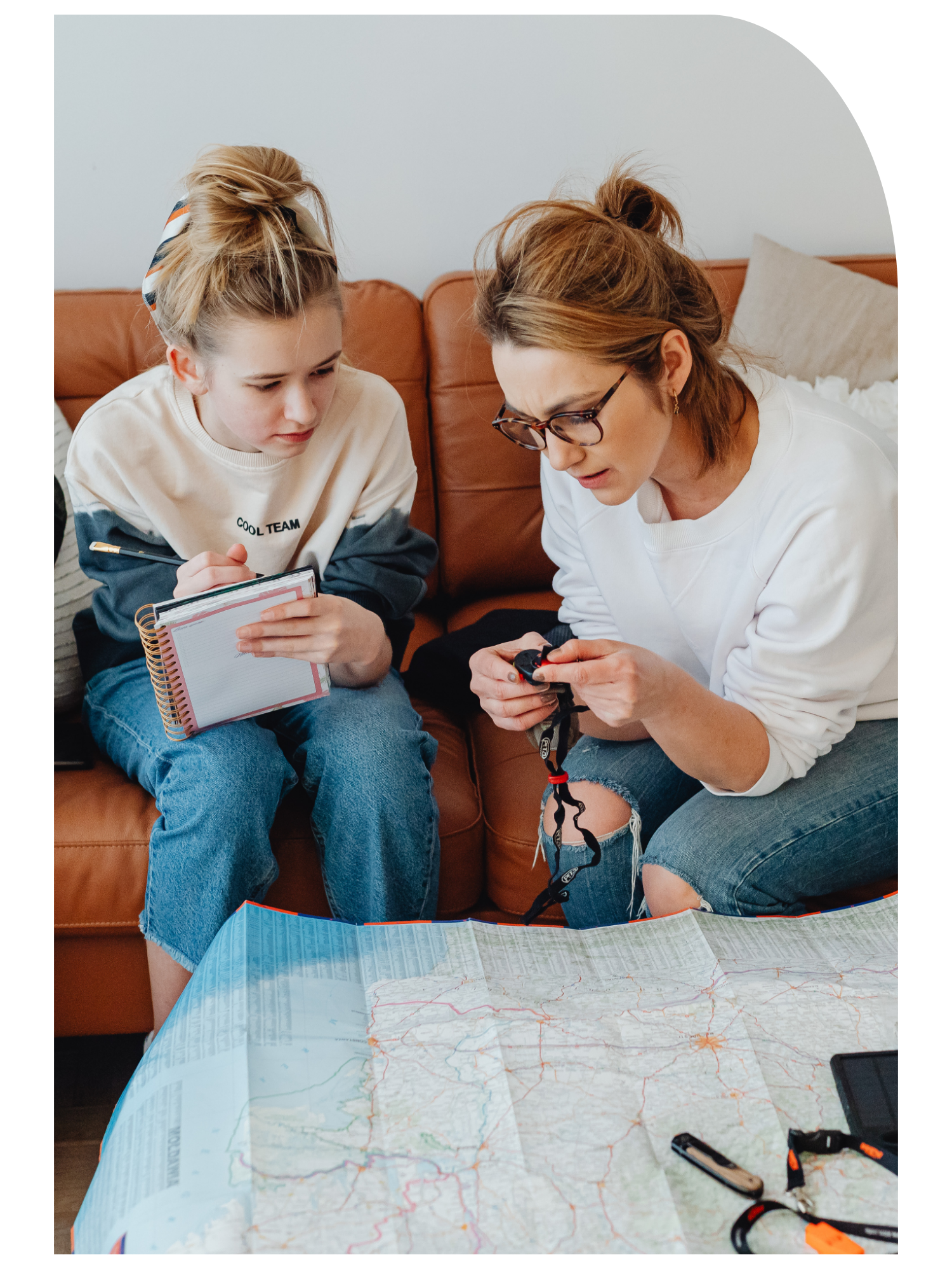 A woman with glasses and a bun hairstyle looking at her phone, sitting on a brown couch with a young girl. A large map with roads and routes is spread out on a table in front of them, with some tools and a black device nearby.