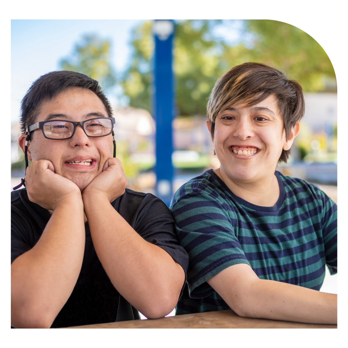 Two young people sitting outdoors, one with glasses and the other smiling, with trees and blue sky in the background.