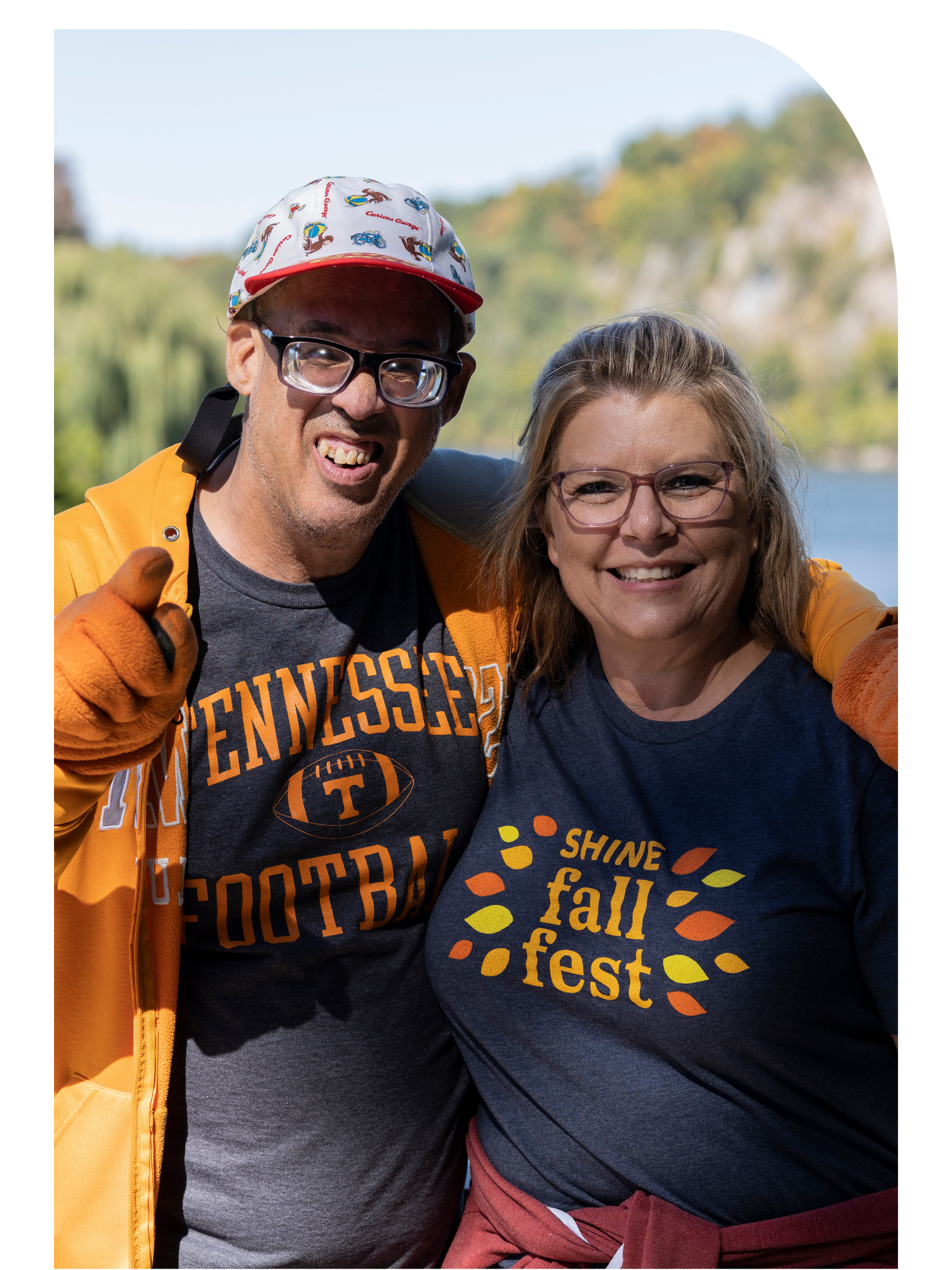 Two smiling people wearing glasses, standing outdoors with trees and blue sky in the background.
