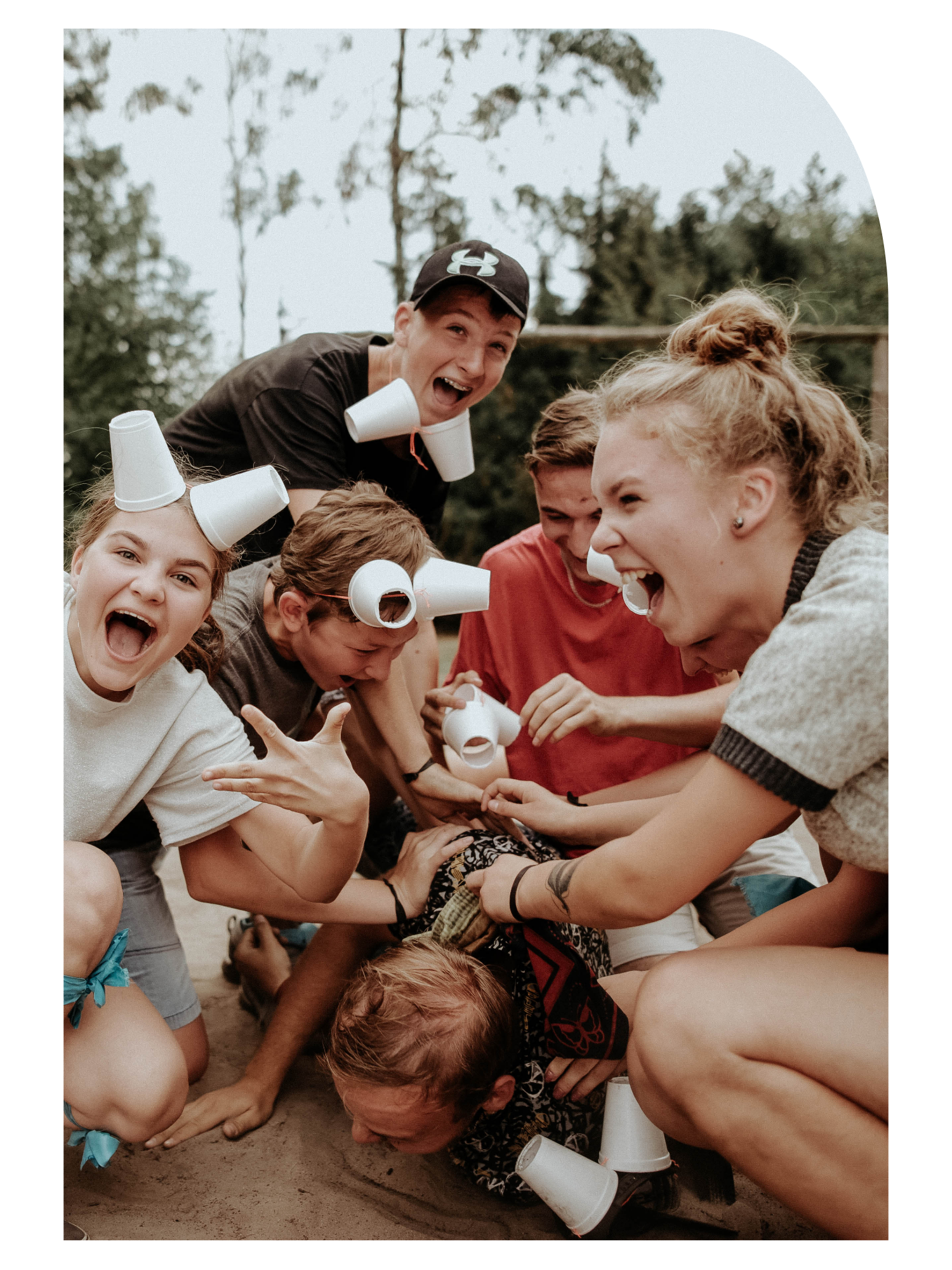 A group of kids and a man engaged in a playful tug-of-war, with cups on their heads, outdoors with trees in the background, on a cloudy day.