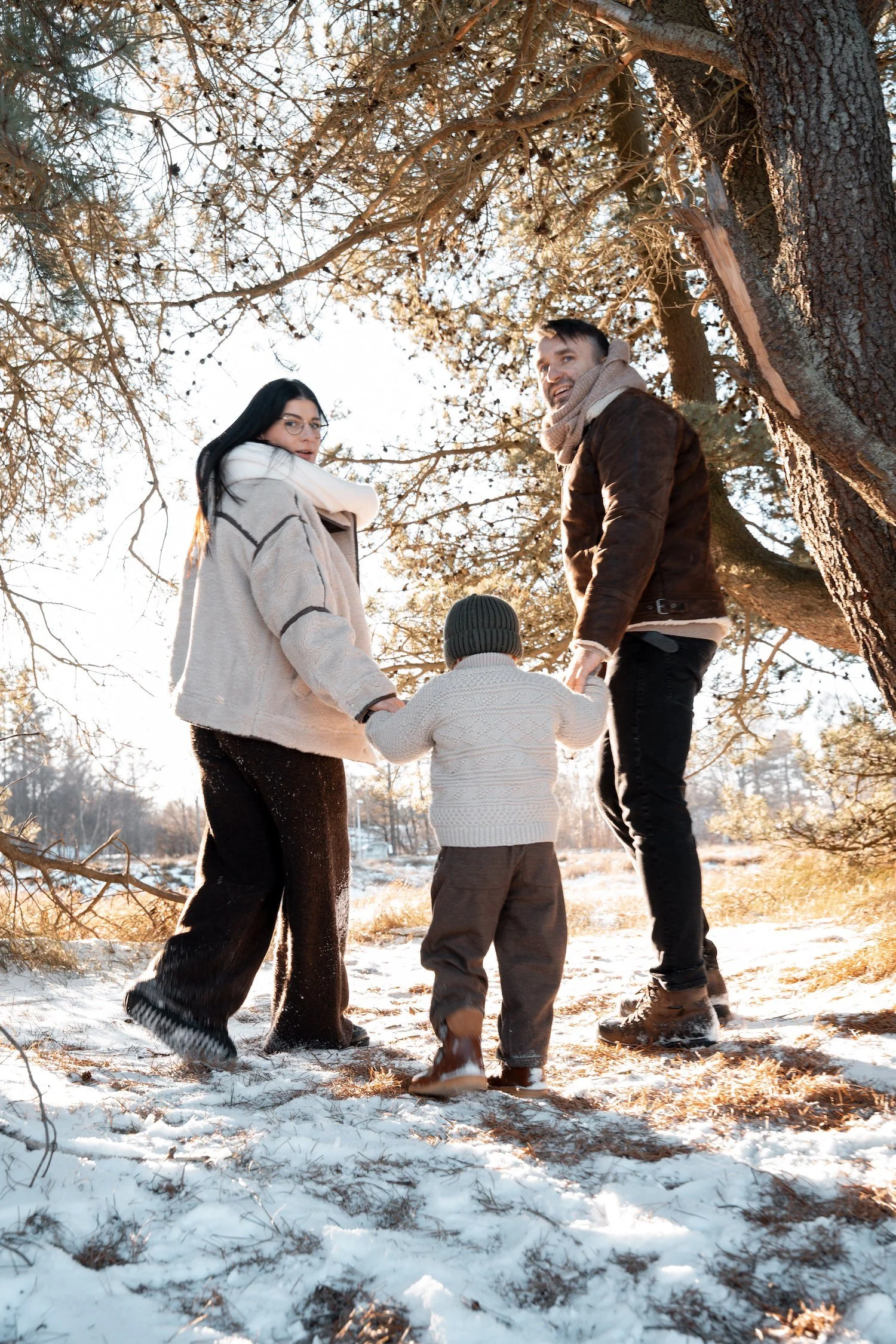familie til fotografering iført vintertøj som er striktrøjer pelsjakker læderjakker strikhuer og læderstøvler. farverne er i beige og brune toner og de går sammen i naturen og kigger på fotograf. Fotograf nordsjælland københavn dyrehaven lyngby