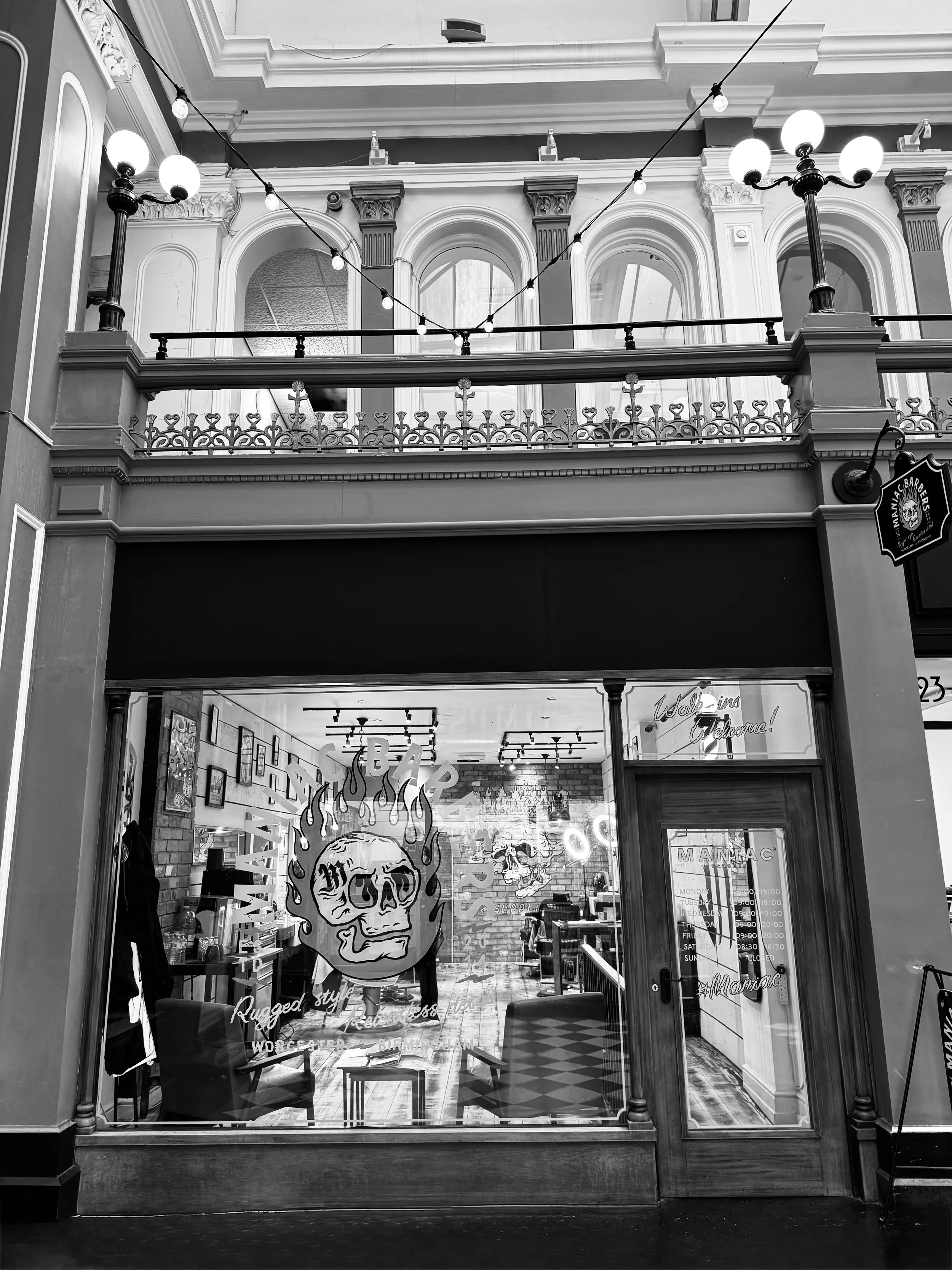 Black and white photo of a barbershop with a large skull graphic with flames on the window, chairs inside, and a sign on the door listing opening hours.