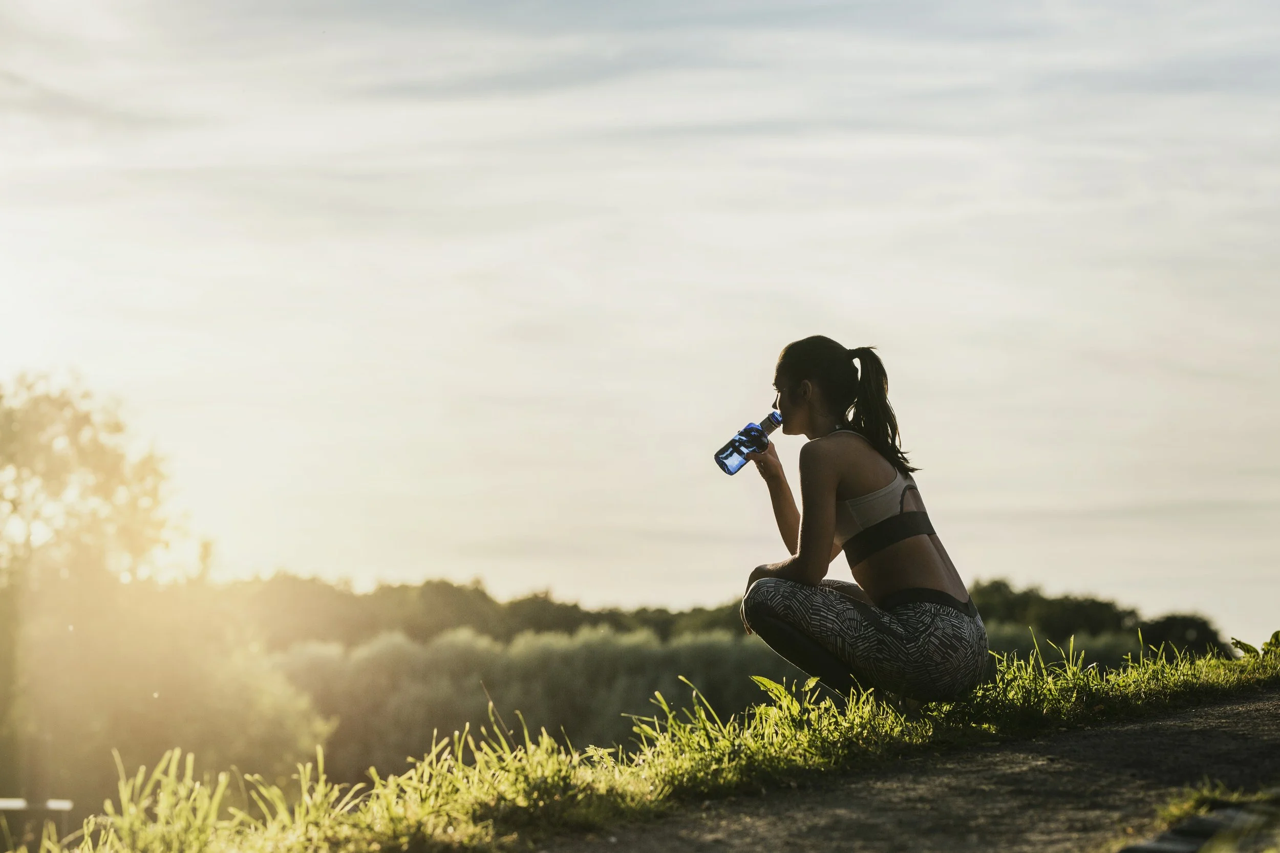A woman in athletic wear is kneeling on the grass near a body of water, drinking from a blue bottle, with a sunset or sunrise in the background.