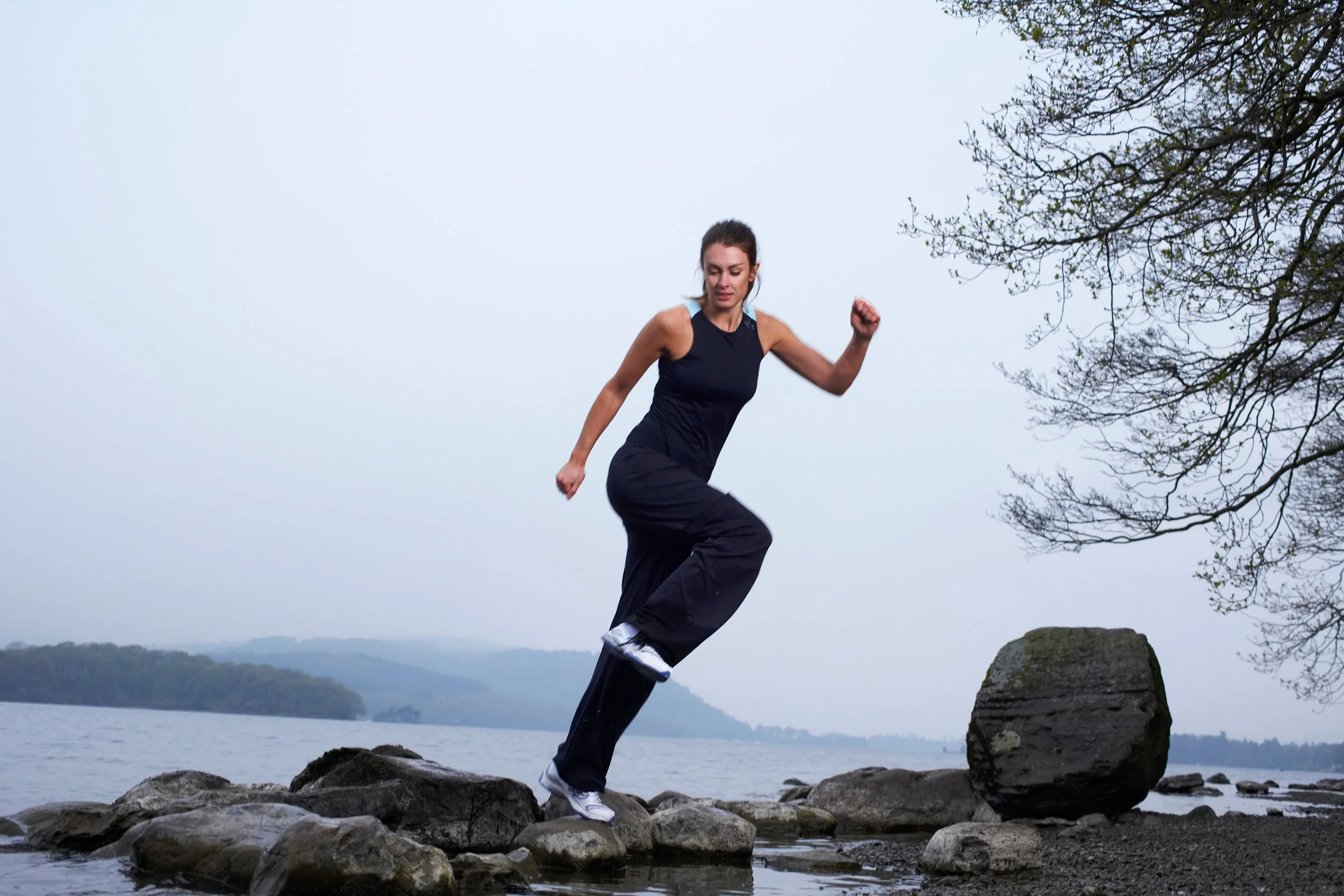 A woman in athletic clothing balancing on rocks near a body of water with trees on the right side and hilly landscape in the background.