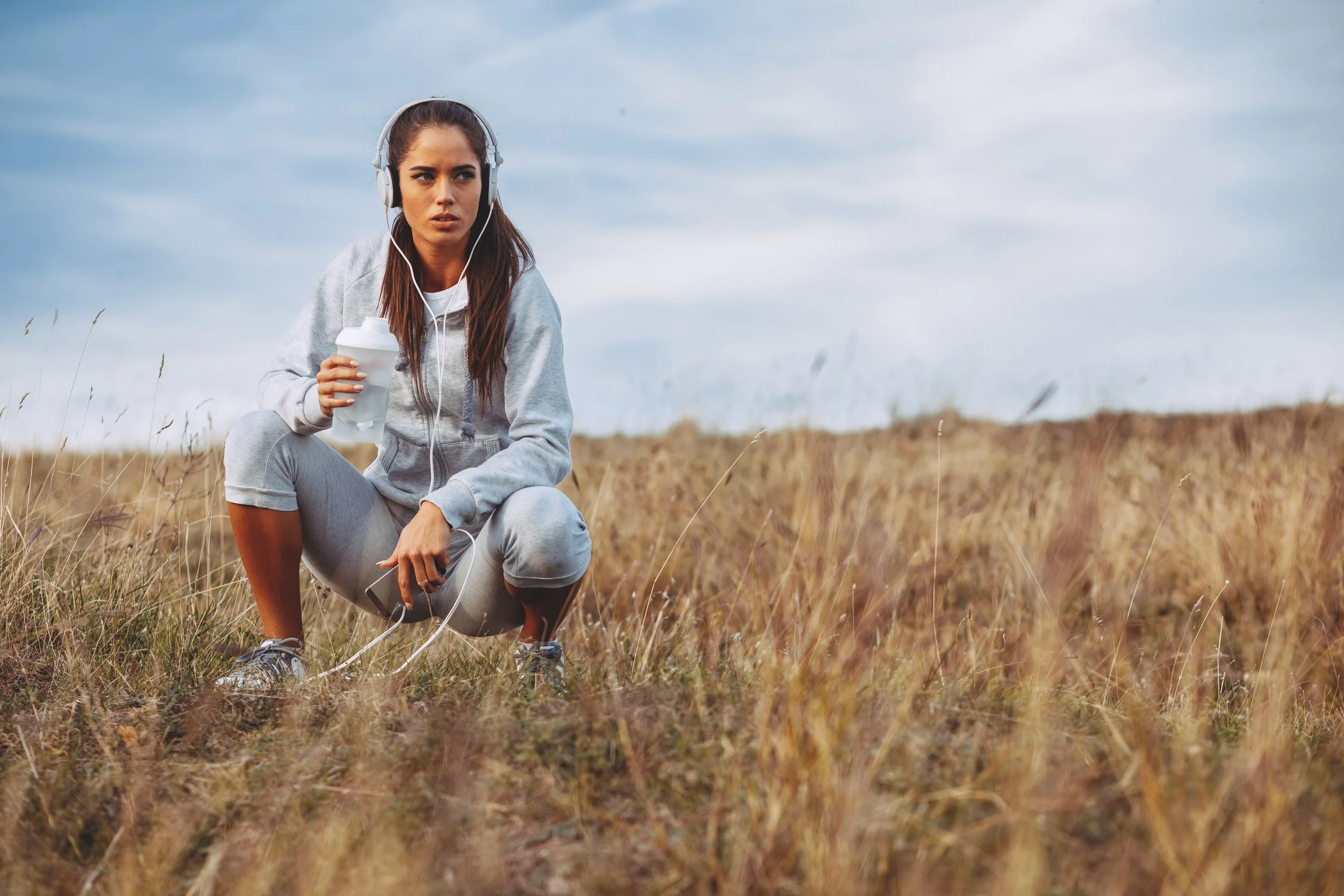 A young woman in athletic clothing squatting in a field, holding a water bottle, listening to music with headphones, with a cloudy sky above.