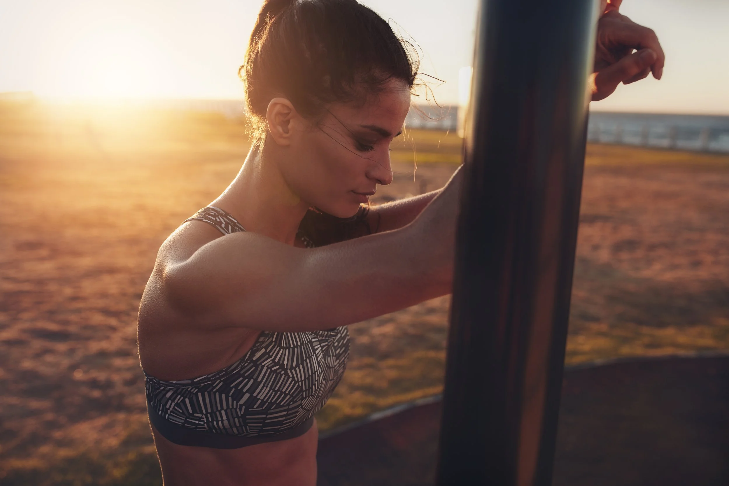 A woman in athletic wear stretching her arms outdoors at sunset near a fence.