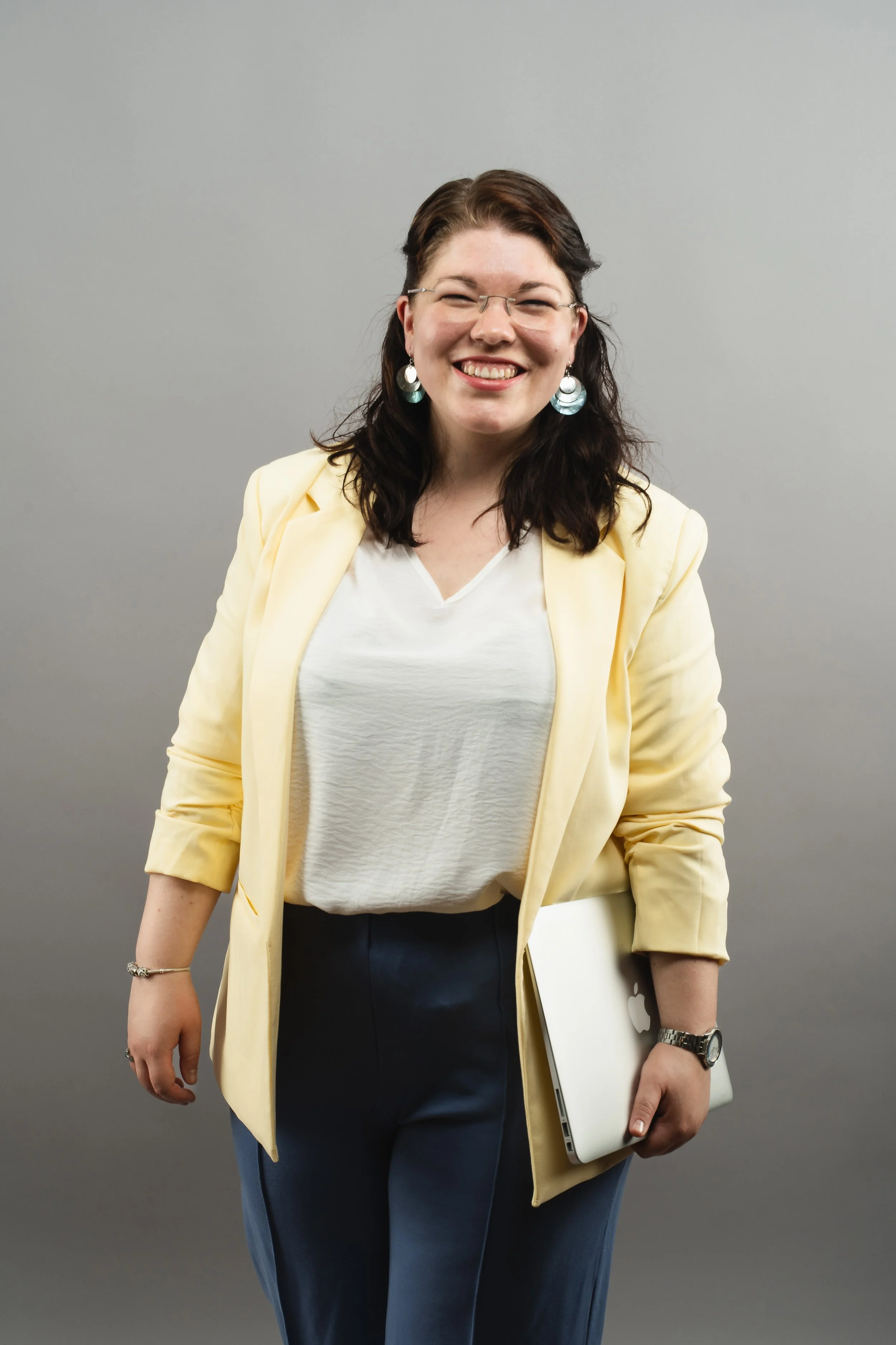 Eine herzlich lachende Frau mit Brille im gelben Blazer steht vor einem grauen Hintergrund. Sie hält einen geschlossenen Laptop locker unter dem Arm und trägt eine blaue Hose, was Professionalität und Lebensfreude zugleich ausstrahlt.