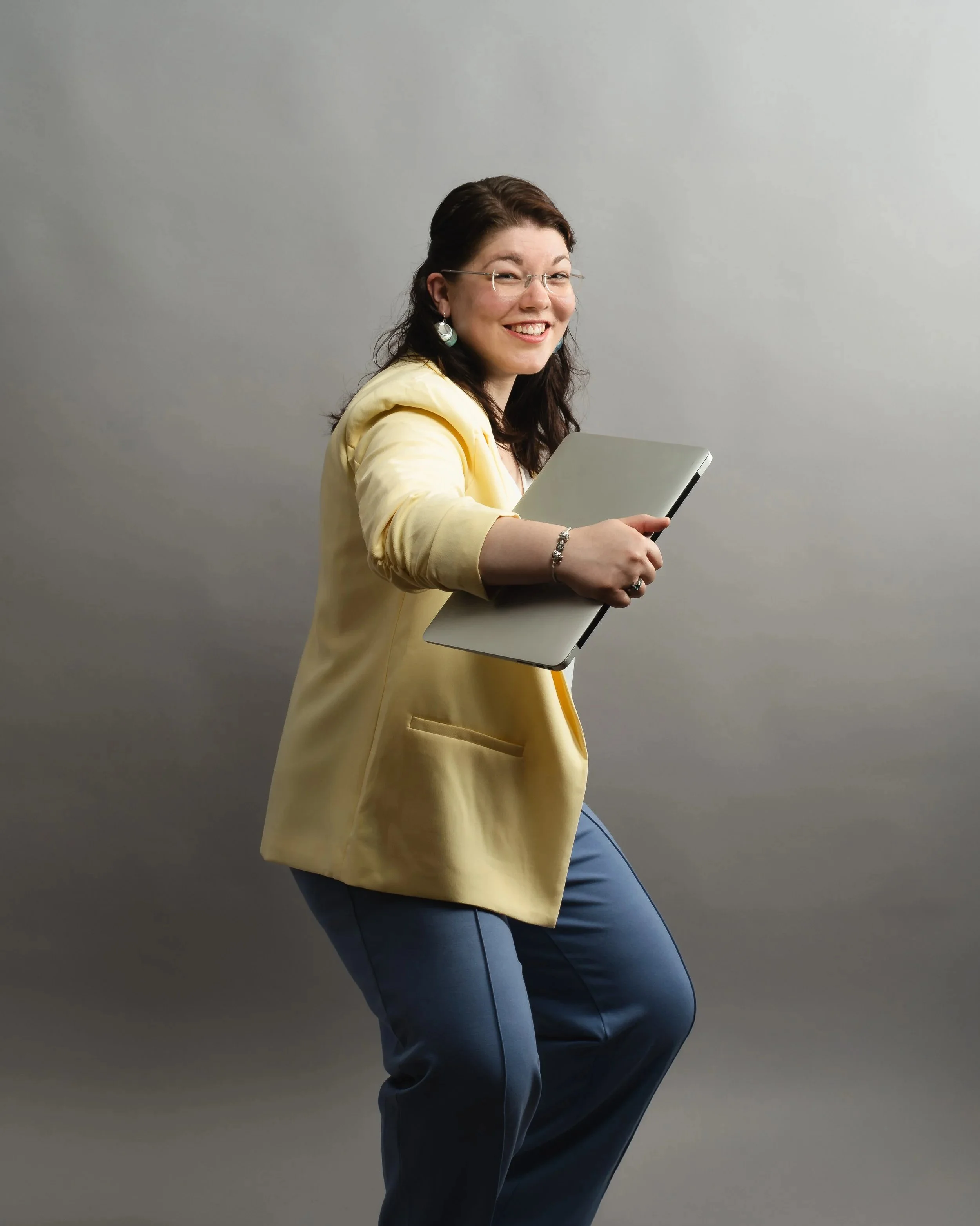 Eine lachende Frau mit Brille im gelben Blazer und blauer Hose macht eine dynamische, fast tänzerische Vorwärtsbewegung. Sie hält einen geschlossenen Laptop wie ein wertvolles Objekt fest umschlungen und blickt voller Freude in die Kamera.