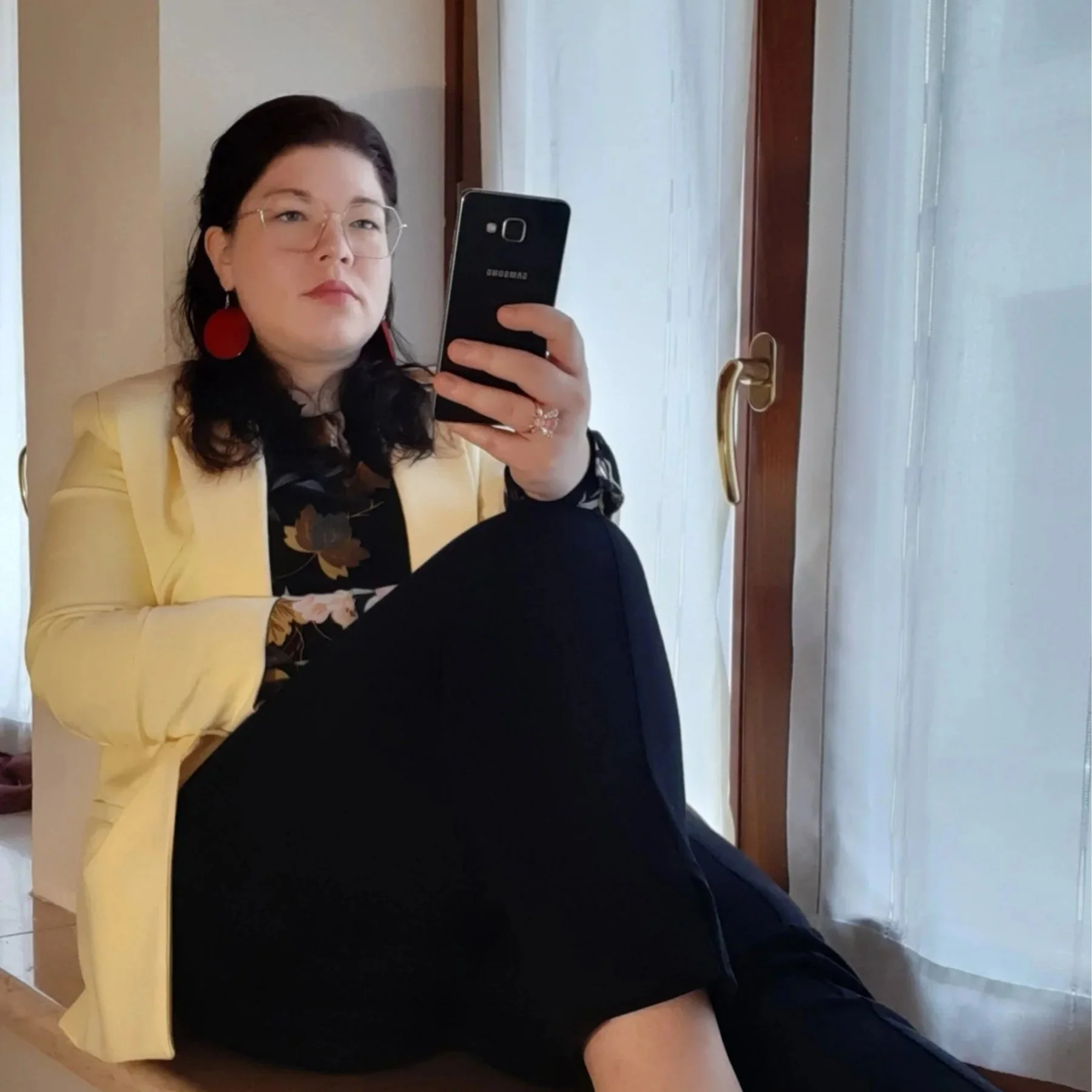 Eine Frau mit Brille und gelbem Blazer sitzt entspannt auf einem Fenstersims und macht ein Selfie. Sie trägt eine dunkle Hose mit Blumenmuster-Bluse und passende gelbe Ballerinas.