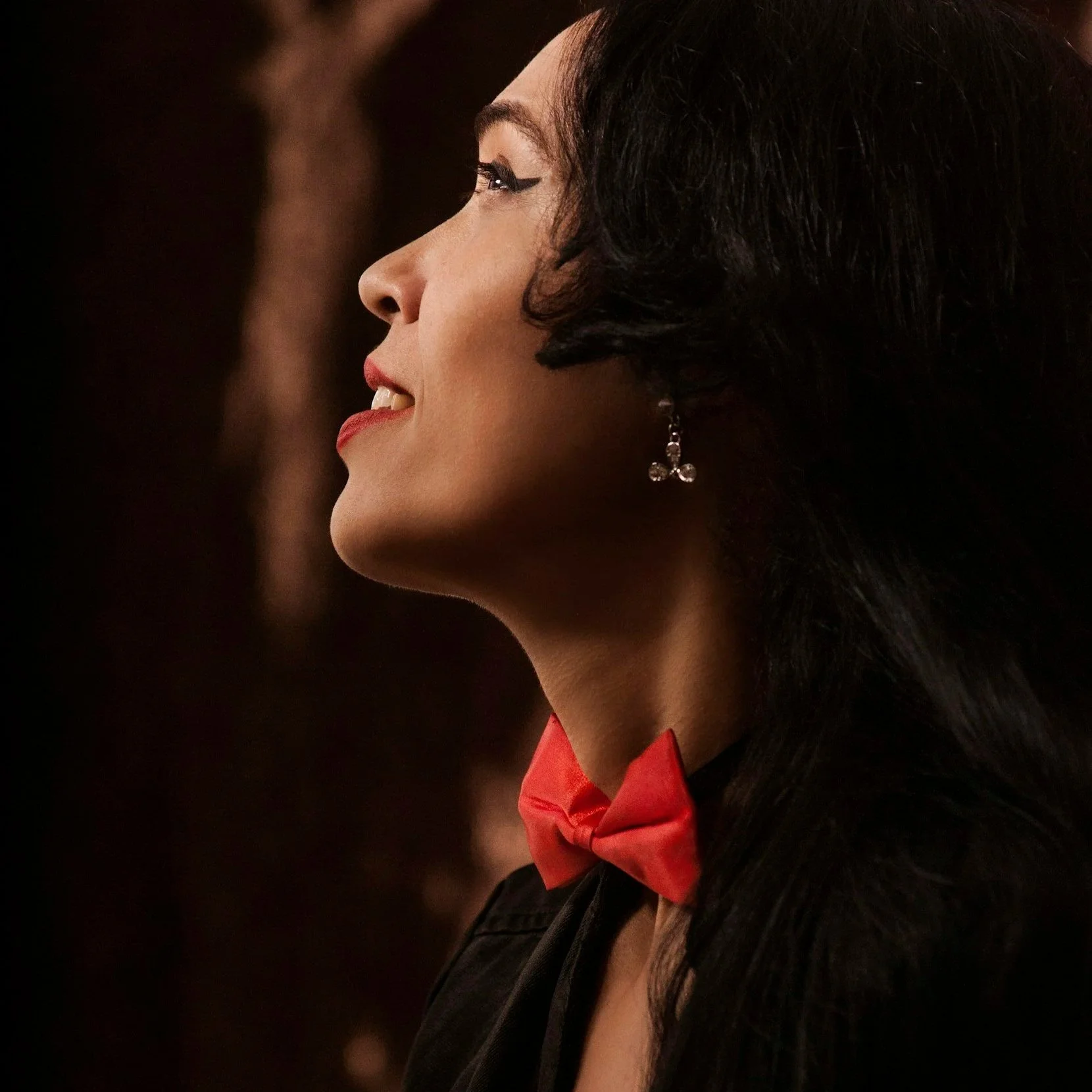 Seitenprofil einer Künstlerin mit dunklem Haar, die mit einem hoffnungsvollen Lächeln nach oben blickt. Sie trägt eine markante rote Fliege und filigrane Ohrringe vor einem dunklen, stimmungsvollen Hintergrund.