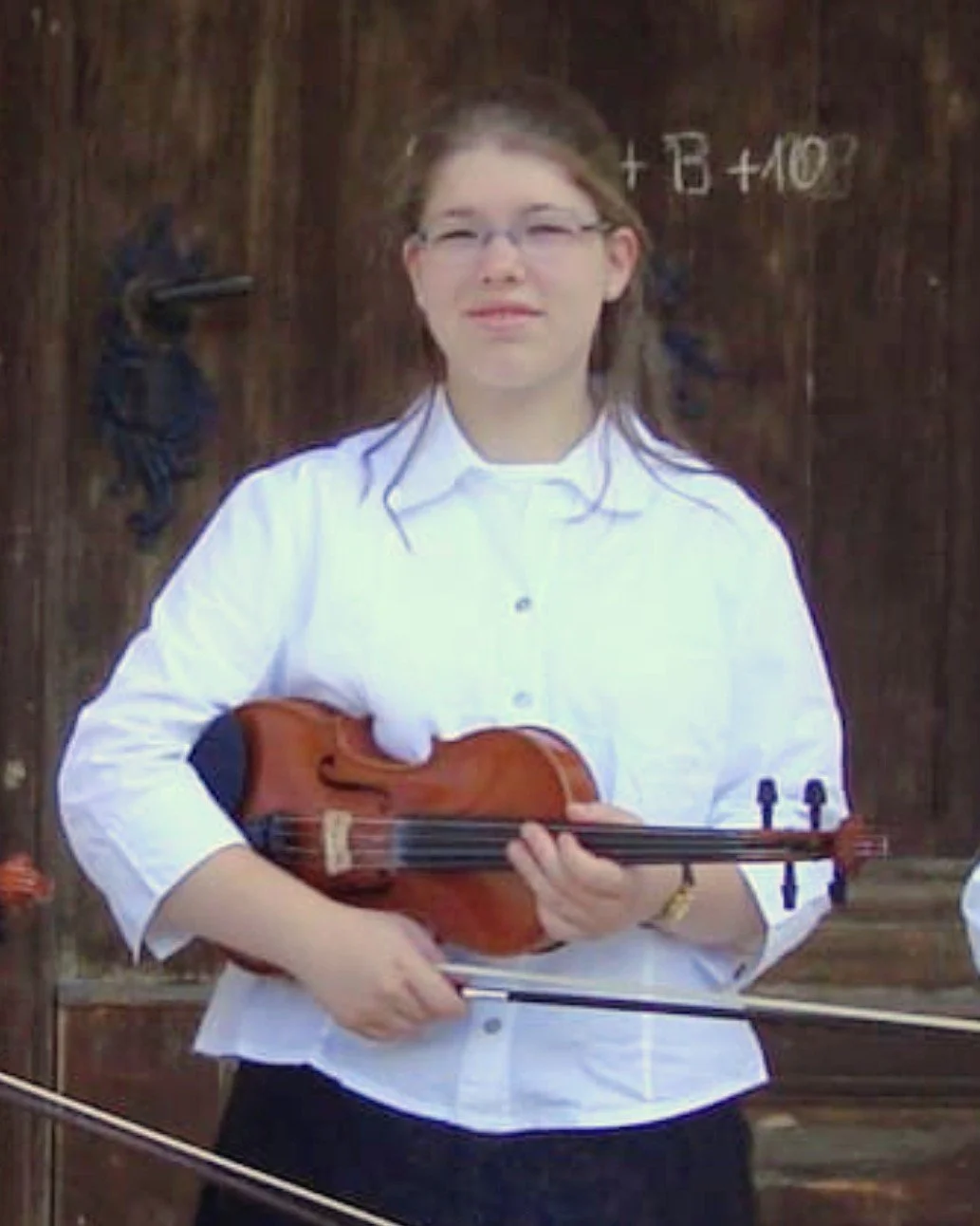 Eine junge Frau mit Brille und weißer Bluse hält eine Violine und einen Bogen vor einer dunklen Holztür.