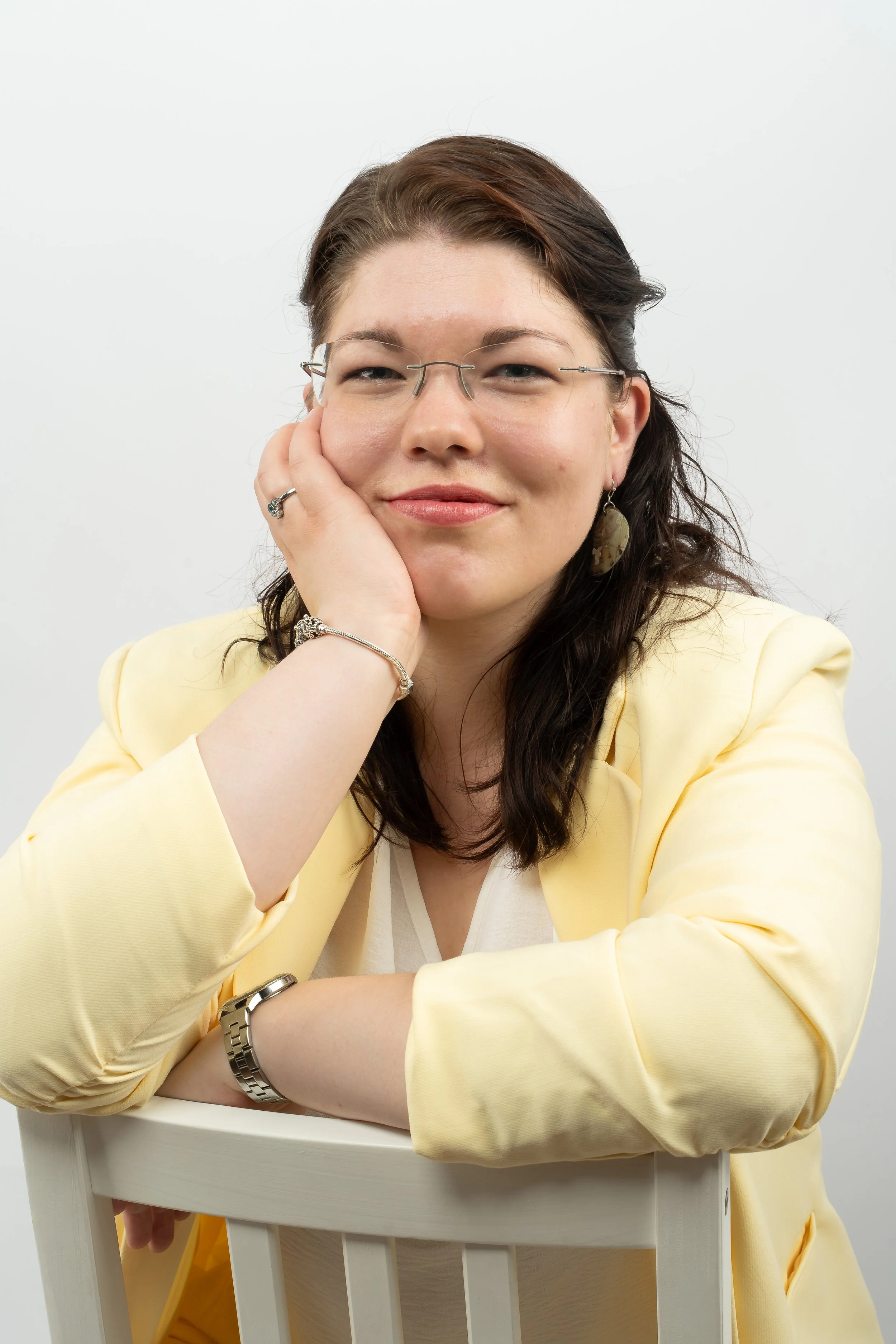 Nahaufnahme einer freundlich lächelnden Frau mit Brille und dunkelbraunem Haar im hellgelben Blazer. Sie stützt ihr Kinn sanft auf die Hand und blickt direkt in die Kamera, was eine vertrauensvolle Atmosphäre schafft.