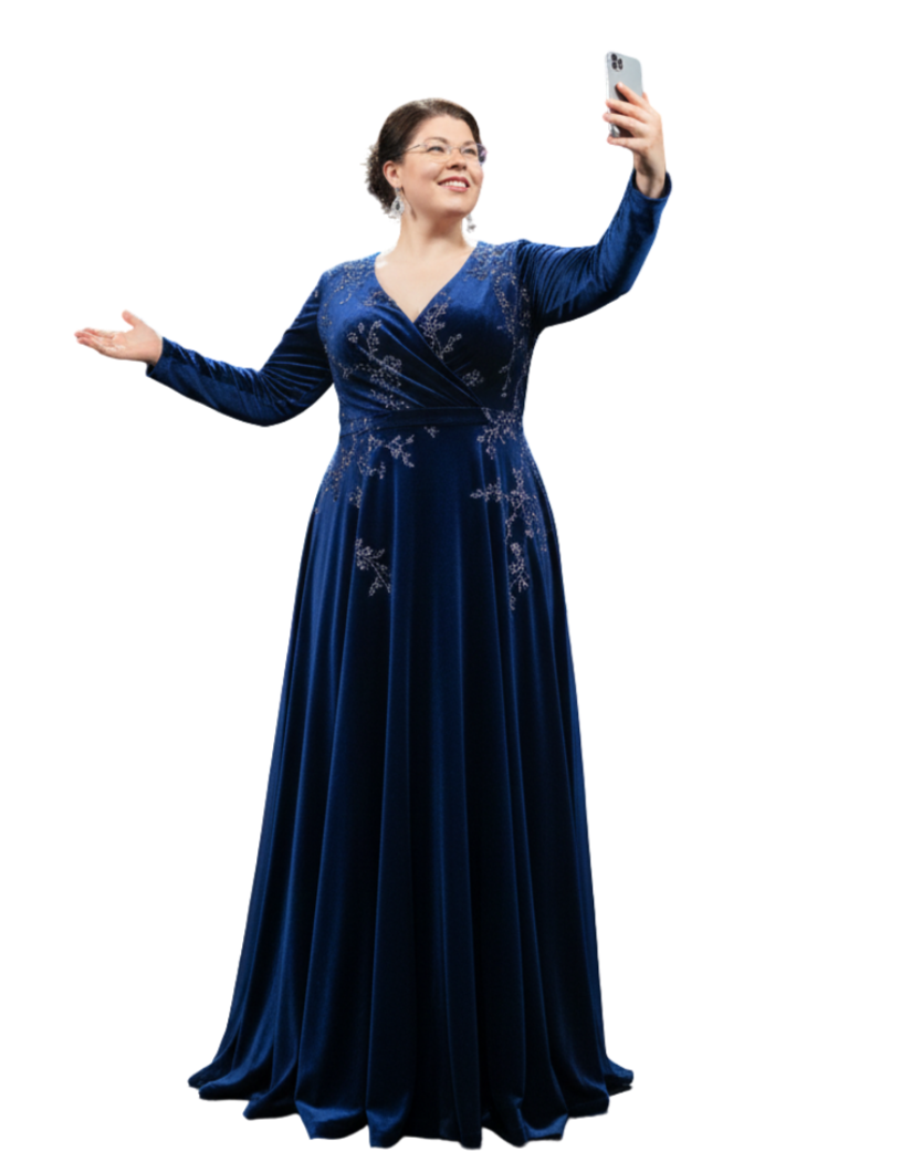 Eine Frau in einem eleganten, bodenlangen dunkelblauen Samtkleid mit silbernen Bestickungen macht lächelnd ein Selfie. Sie hält das Smartphone hoch und öffnet den anderen Arm einladend zur Seite vor weißem Hintergrund.