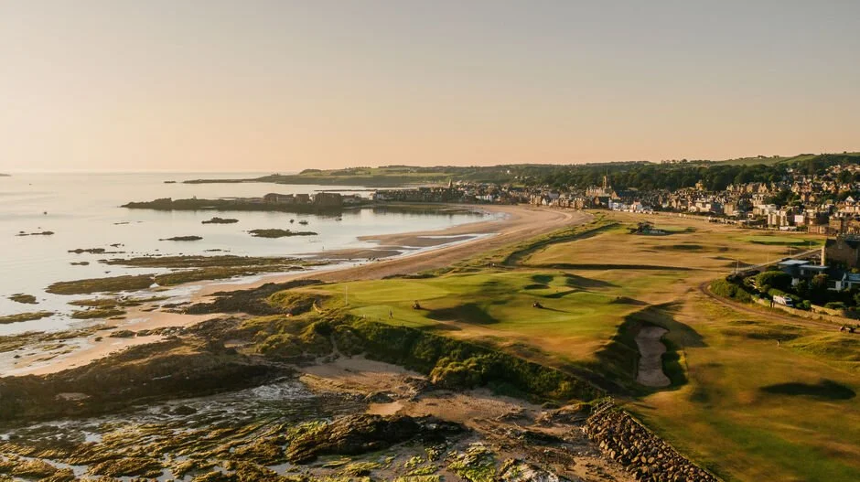 A coastal golf course with green fairways and sand traps, overlooking a beach and rocky shoreline, with a town in the background under a clear sky. St andrews, Kingsbarns.
