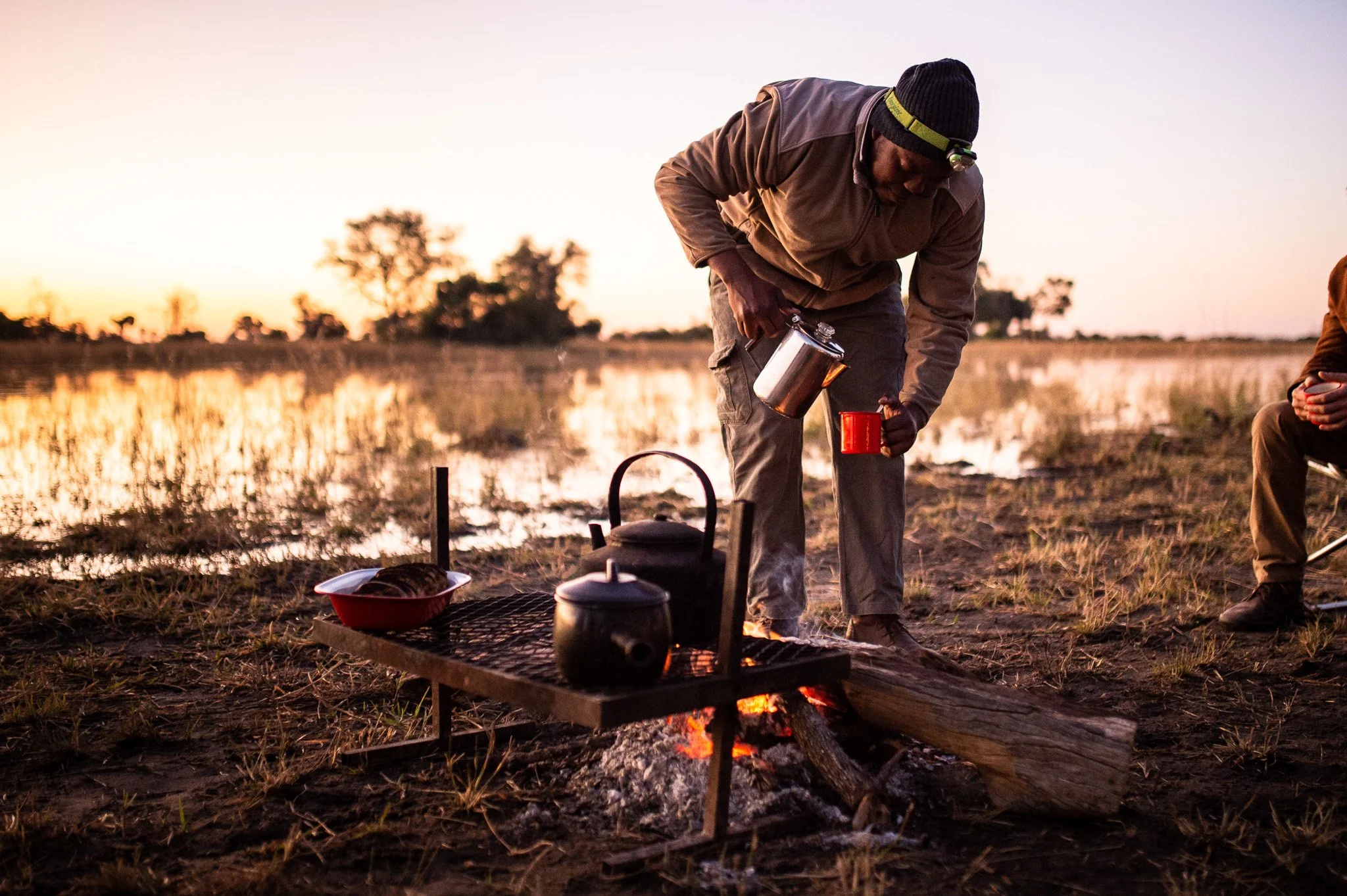 A person pouring coffee into a mug near a campfire at sunset by a lake, with trees in the background.