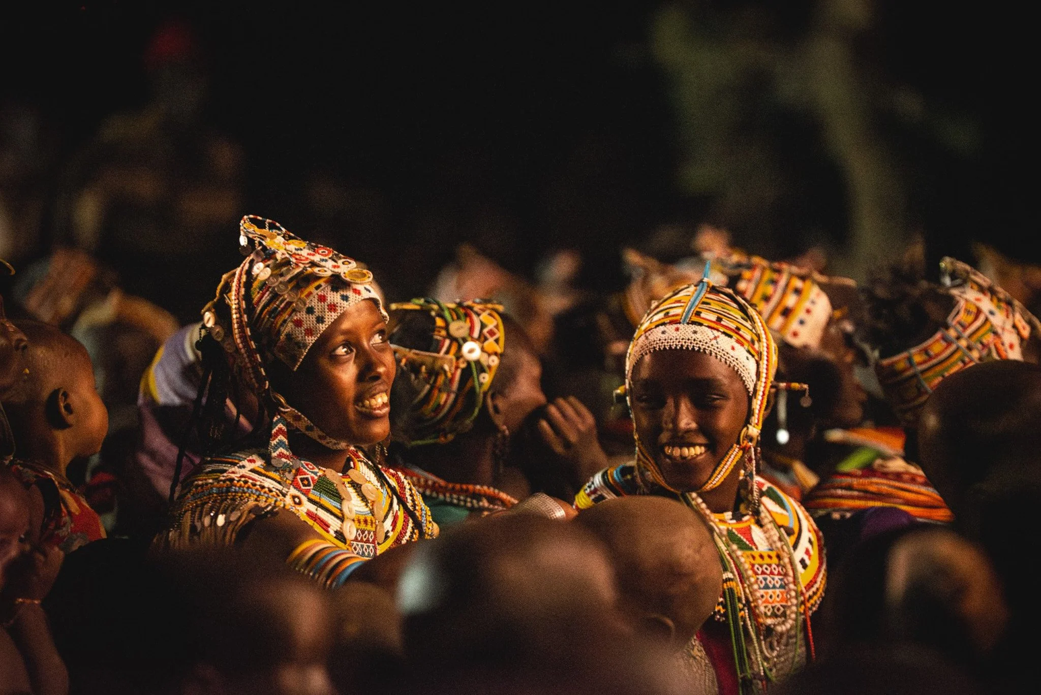 Group of women in traditional colorful attire and headpieces, gathered outdoors at night, smiling and engaging with each other.