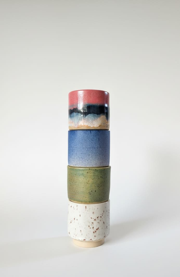 Four stacked ceramic cups with glazed, colorful finishes in pink, black, blue, green, and white speckled, against a plain light background.