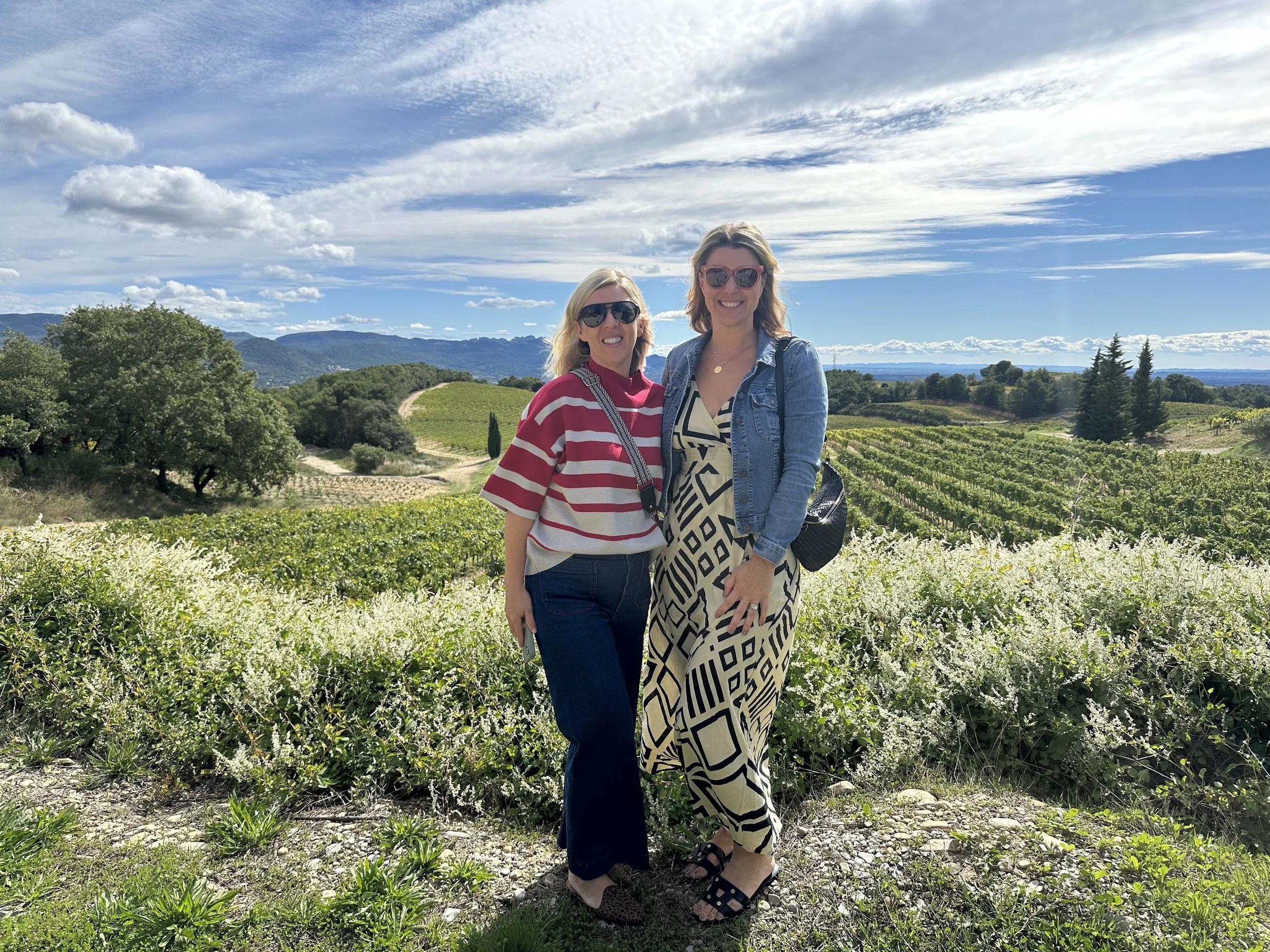 Two women smile and pose for a photo in a vineyard with rolling hills and a partly cloudy sky in the background.
