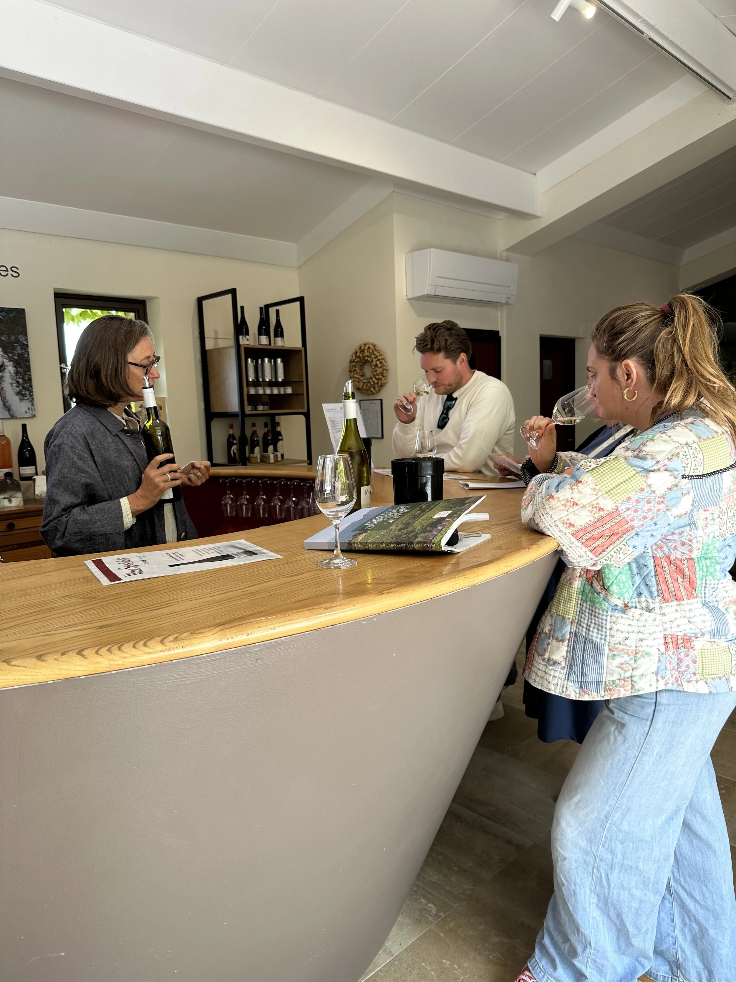 People participating in a wine tasting at a counter in a winery or tasting room, with glasses of wine, a wine bottle, and tasting notes in front of them.