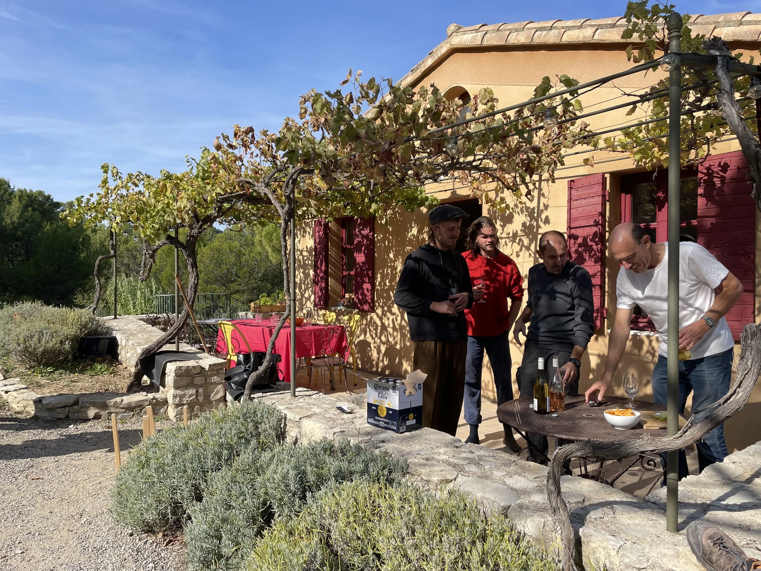A group of five people standing around a small table with drinks and snacks on an outdoor patio with a yellow house that has red shutters and grapevine-covered trellis, surrounded by greenery.