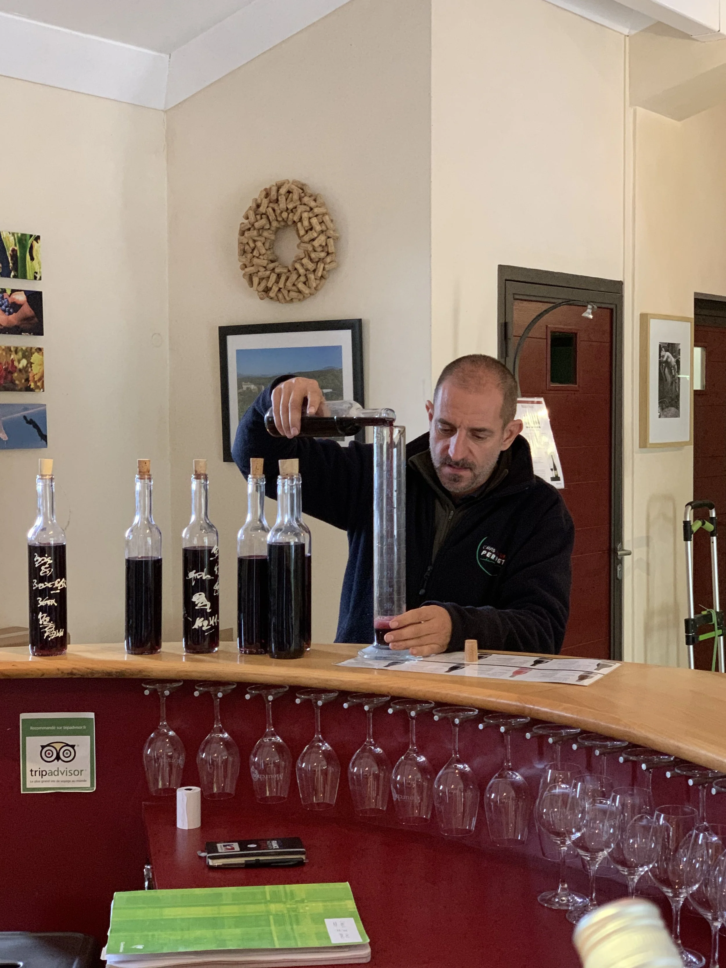A man pouring red wine into a glass at a bar with bottles of wine, glasses, and a small cork on the counter.
