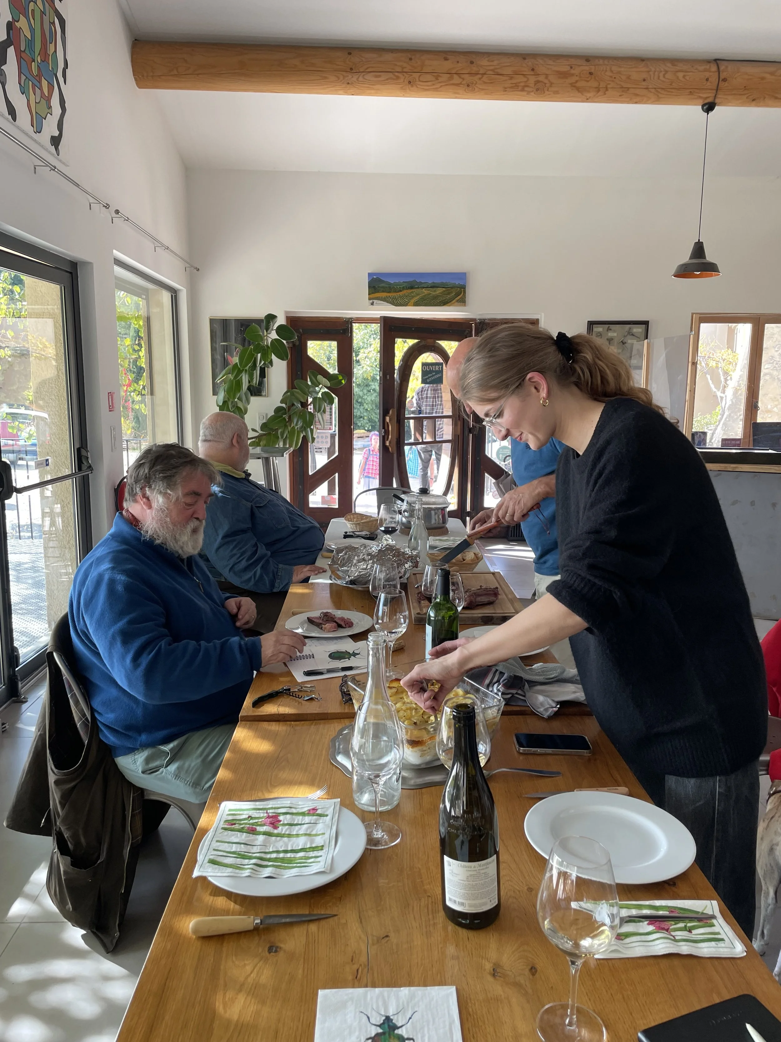 People gathering around a wooden dining table inside a cozy restaurant enjoying a meal, with sunlight coming through large windows.