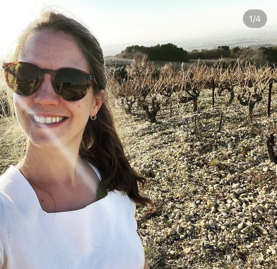 A woman wearing sunglasses and a white shirt is taking a selfie in a vineyard with rows of grapevines, rocky ground, and a distant landscape in the background.