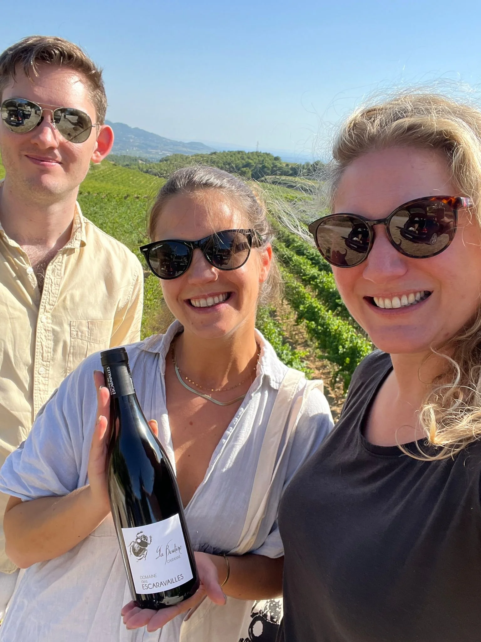 Three people outdoors in a vineyard, smiling and wearing sunglasses, one holding a bottle of wine.