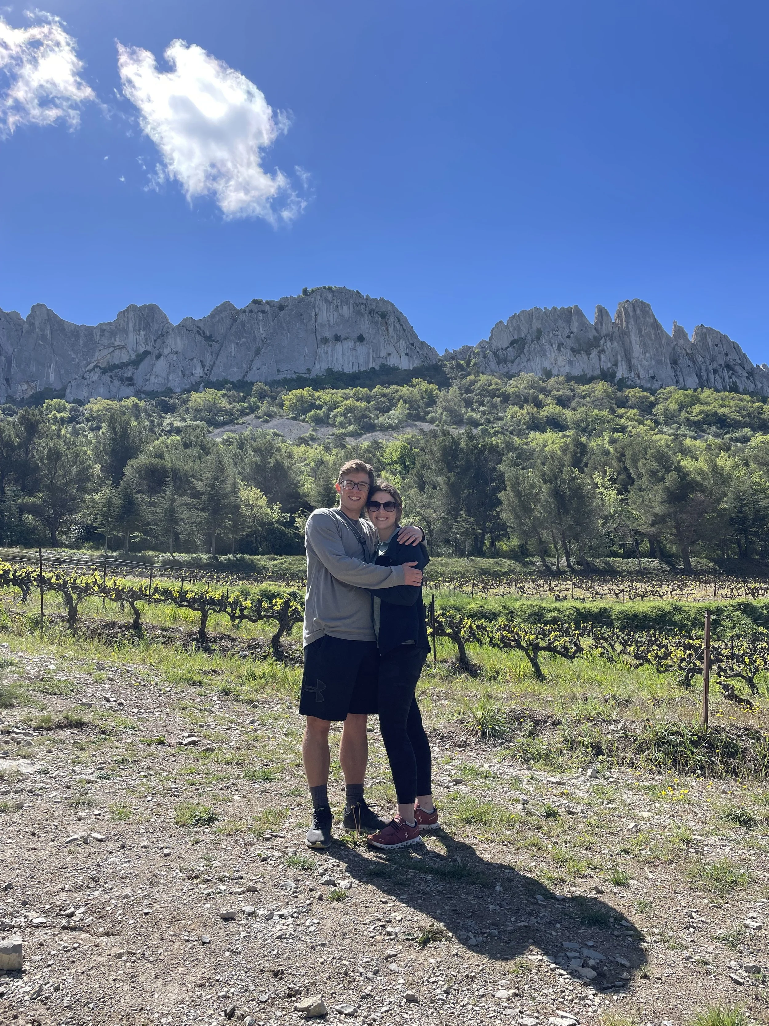 A couple hugging outdoors in a vineyard with a mountain range and a blue sky in the background.