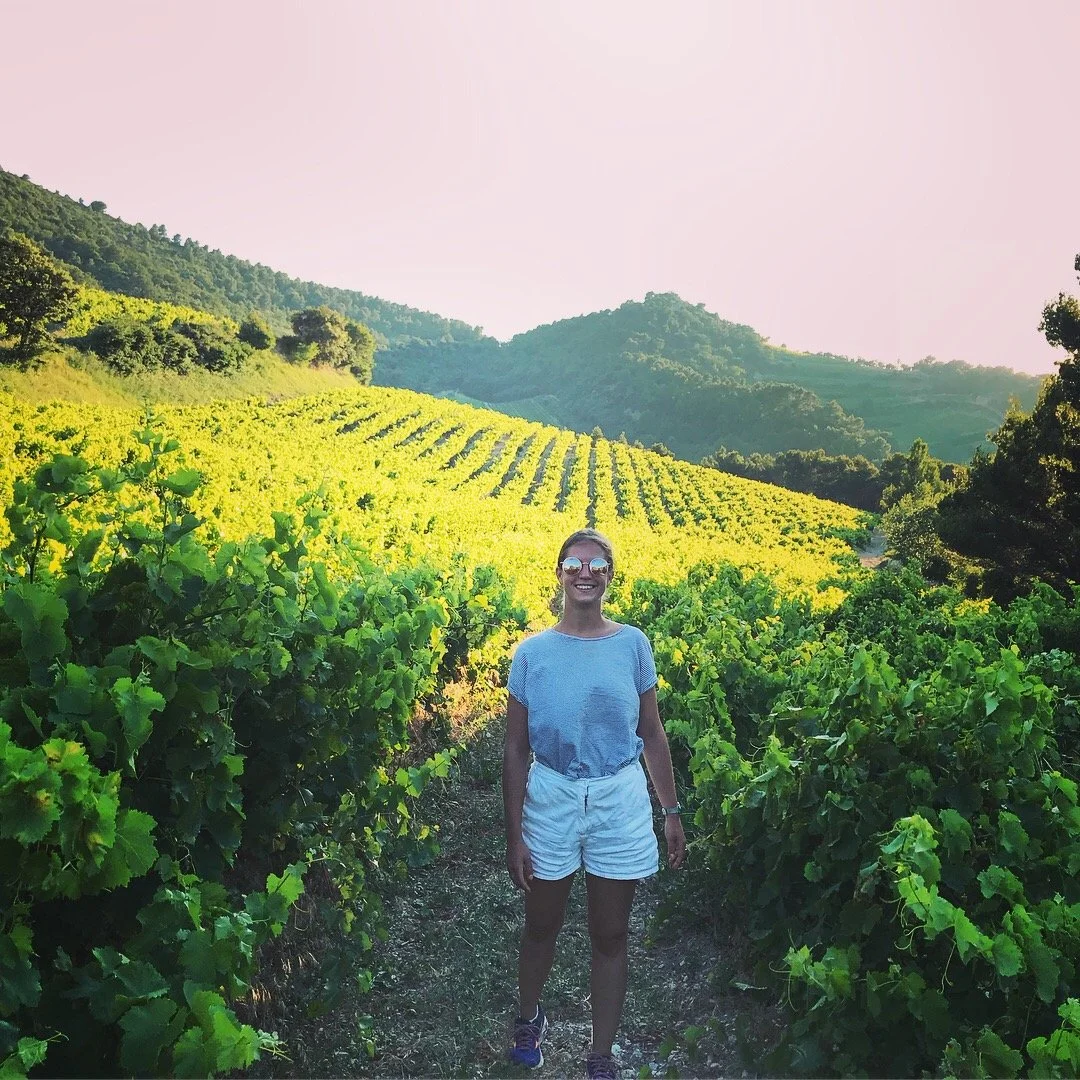 A woman standing on a vineyard path, smiling, with green grapevines and rolling hills in the background on a sunny day.