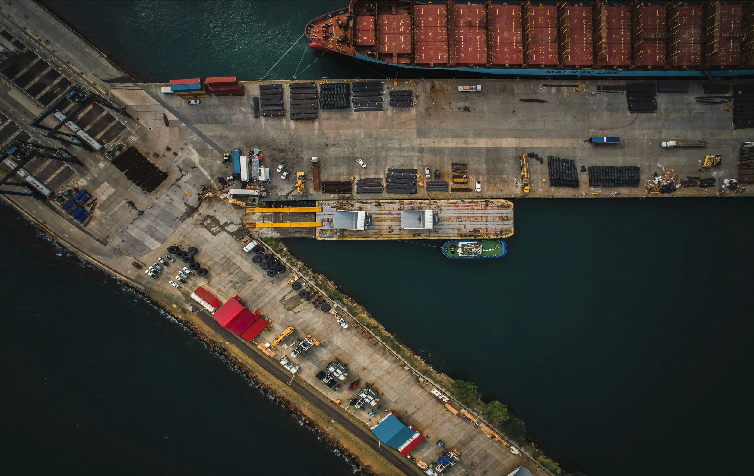 An aerial view of a dock with a cargo ship, containers, trucks, and vehicles along the waterfront.