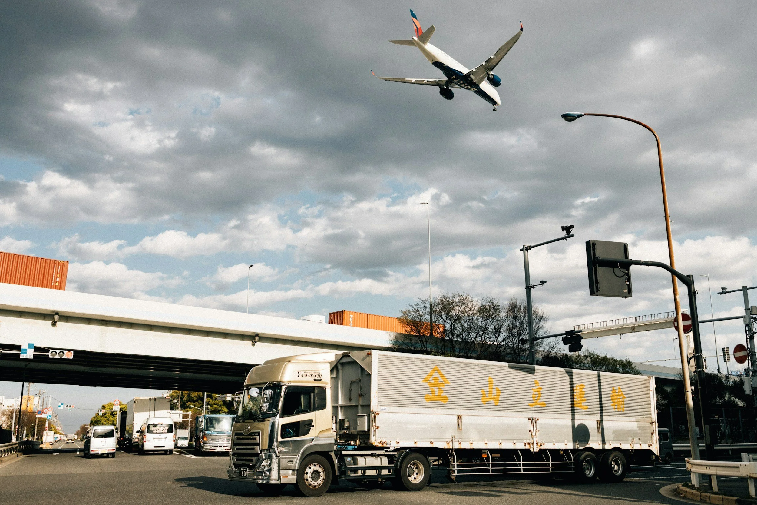 An airplane flying low above a highway, with vehicles and an overpass below. Overcast sky with dark clouds.