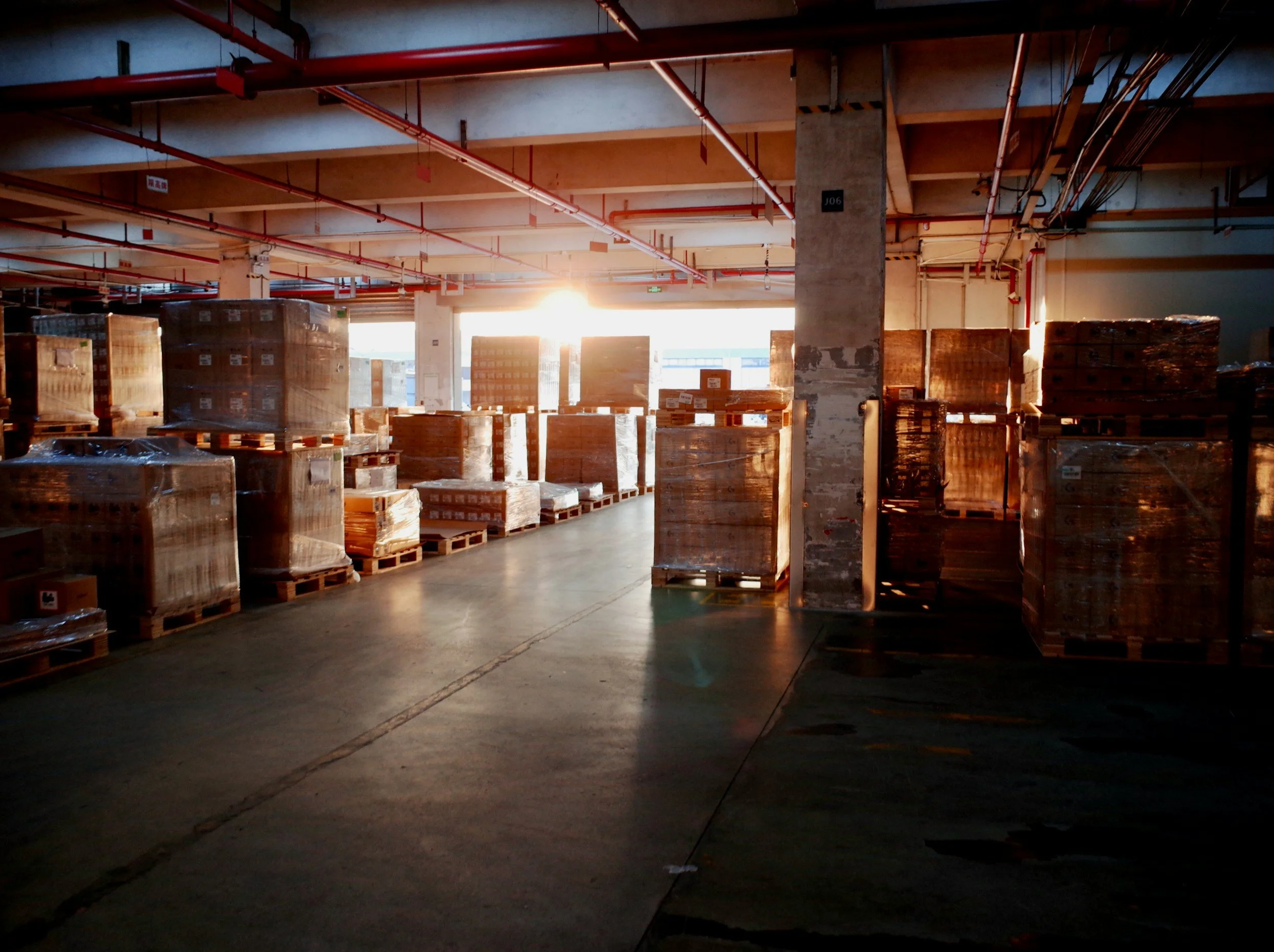 A warehouse with stacks of pallets wrapped in plastic, sunlight coming through large windows, visible pipes and concrete pillars.