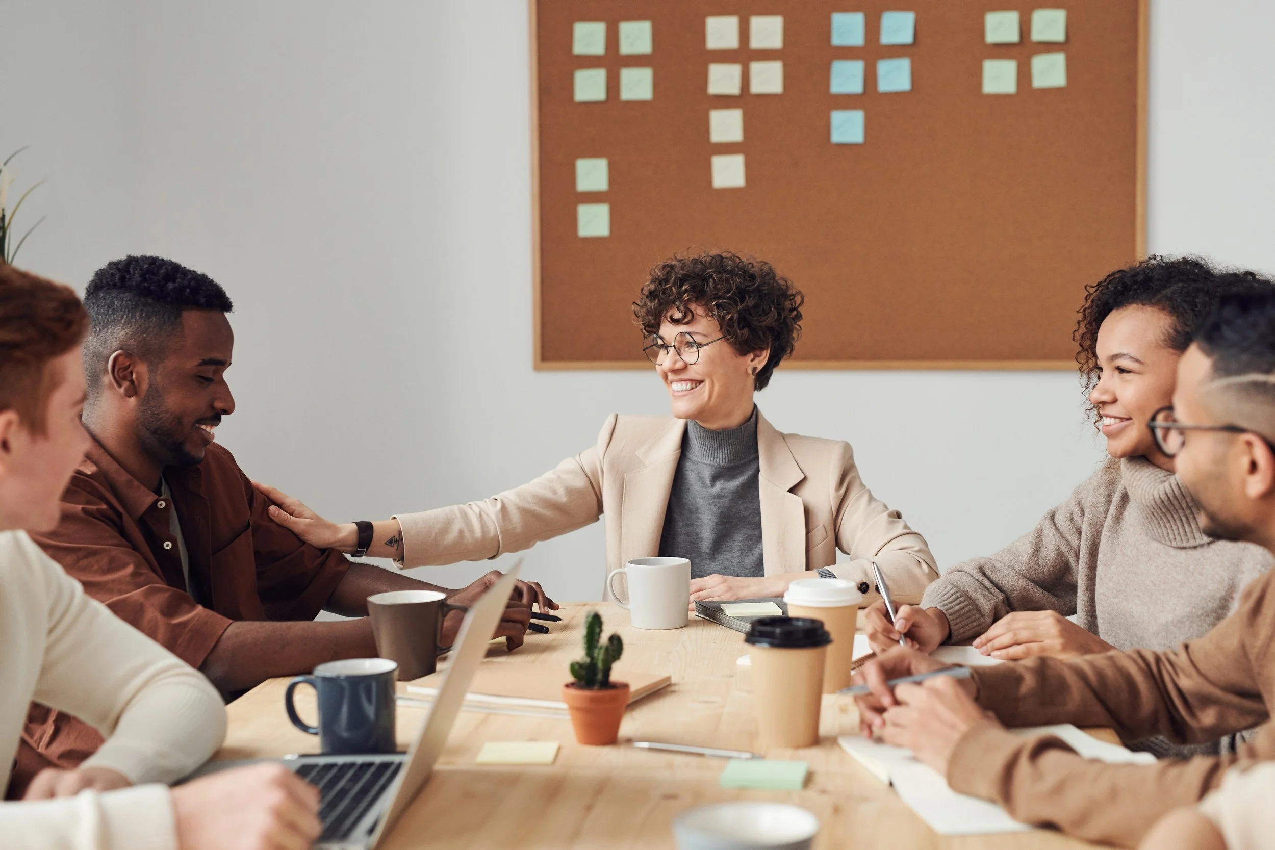 A diverse group of people sitting at a wooden conference table, smiling and engaging in a discussion with a woman in a beige blazer leading the conversation. Several coffee mugs and laptops are on the table, with a corkboard filled with sticky notes on the wall behind them.