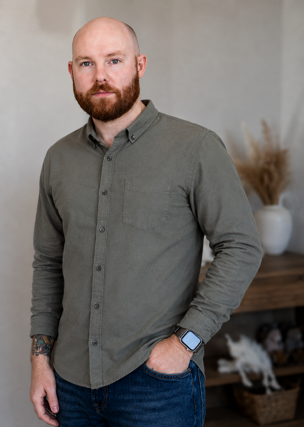 A bald man with a red beard wearing a gray button-up shirt and blue jeans, standing indoors with a neutral background and a decorative vase with dried plants behind him.