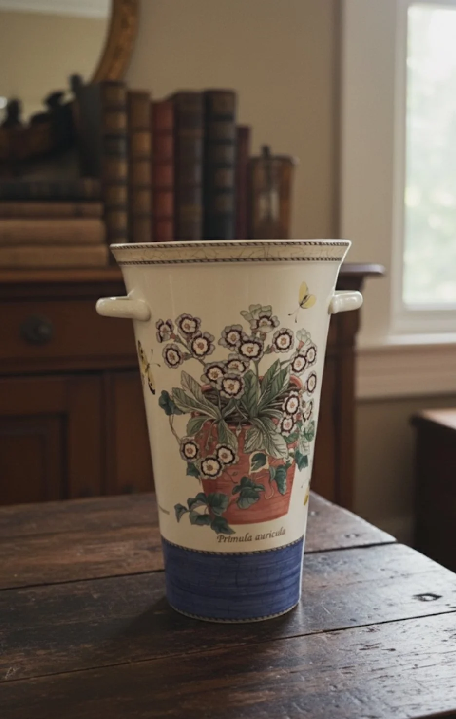 Decorative ceramic vase with floral design and the scientific name 'Primula auricula' on a wooden table, in front of a wooden cabinet with books and a mirror, near a window.