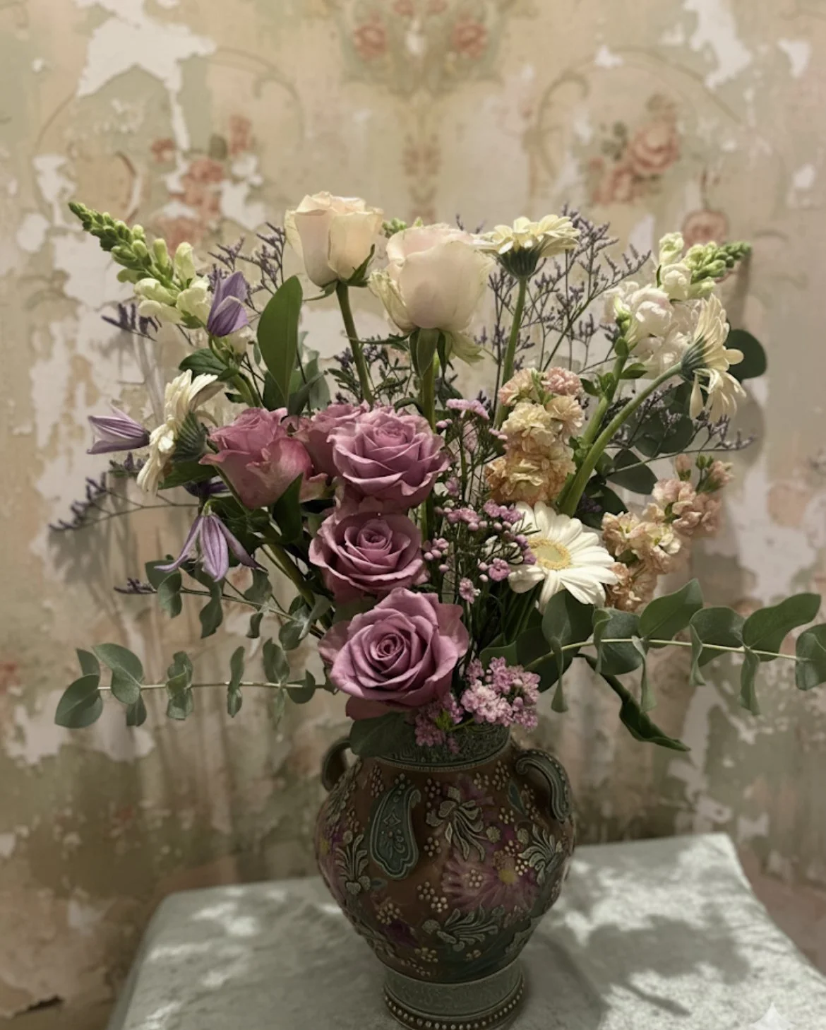 A decorative vase filled with a bouquet of pink roses, white daisies, lilac-colored flowers, and various greenery, placed on a white surface in front of a textured, painting-like background.