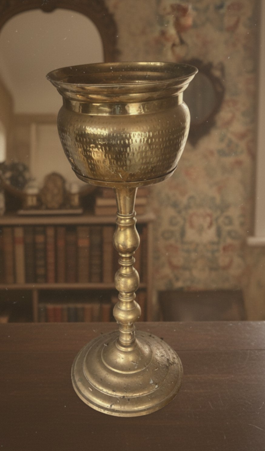 A gold-colored ornate goblet with a textured bowl, a decorated stem, and a round base, sitting on a dark wooden surface in a room with bookshelves and decorative objects in the background.